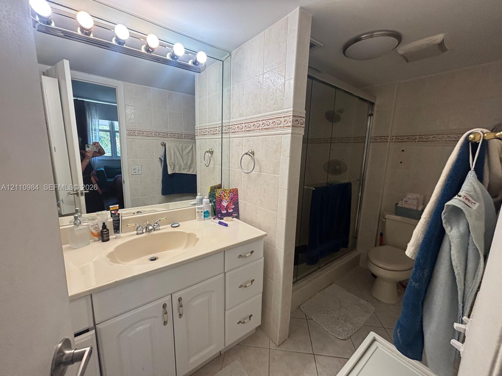 Bathroom featuring a white vanity cabinet with an integrated sink, a large wall-mounted mirror with an overhead five-light fixture, ceramic wall tiling with a decorative border, and a glass-enclosed shower