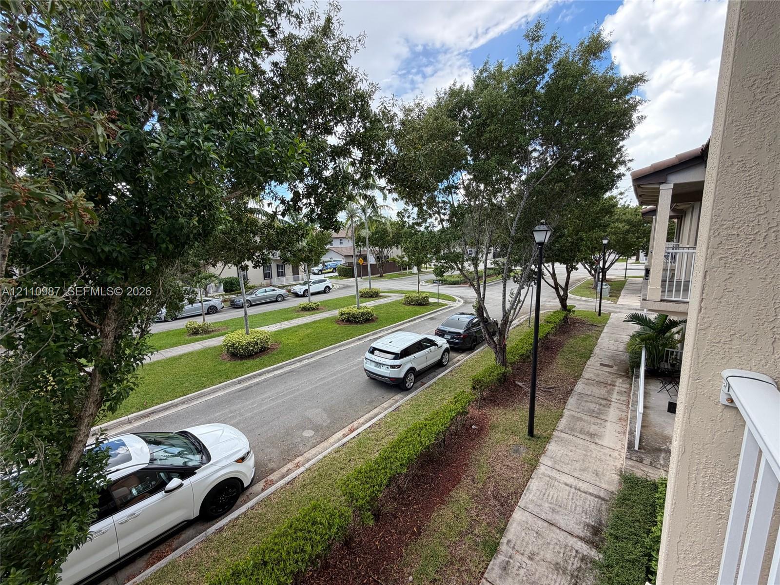 Exterior streetscape featuring a paved walkway, mature landscaping, manicured grass medians, and post-mounted outdoor lighting