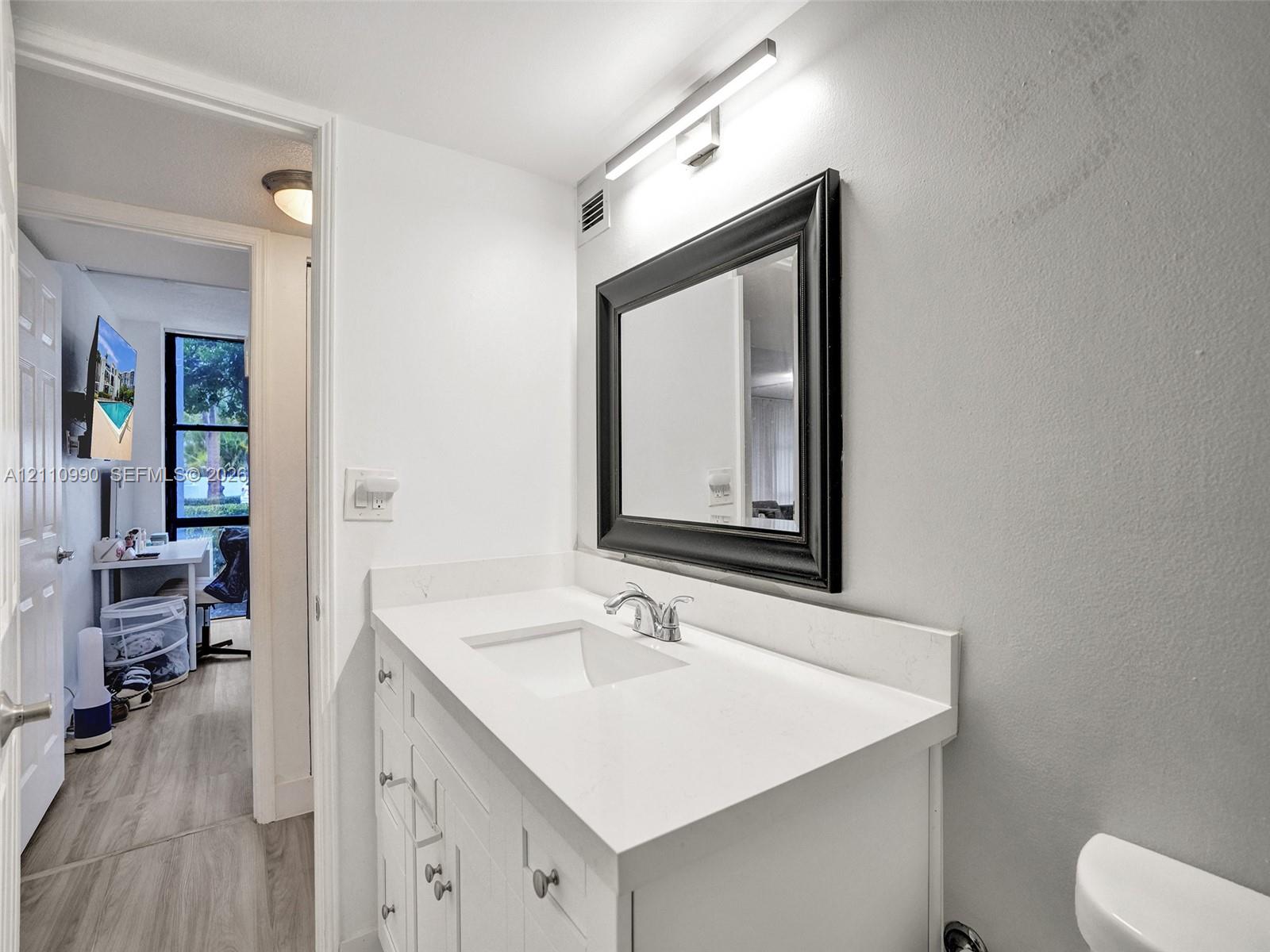 White vanity with a solid-surface countertop and integrated sink, complemented by a framed mirror, wall-mounted vanity lighting, and matte-finish hardware