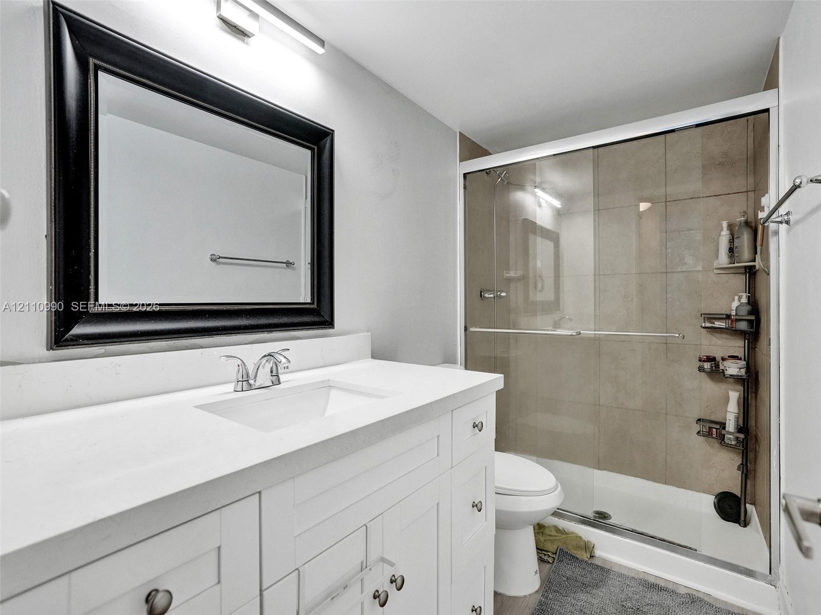 White shaker-style vanity with a solid surface countertop, rectangular integrated sink, chrome faucet, framed mirror, and tiled shower enclosure with a glass sliding door