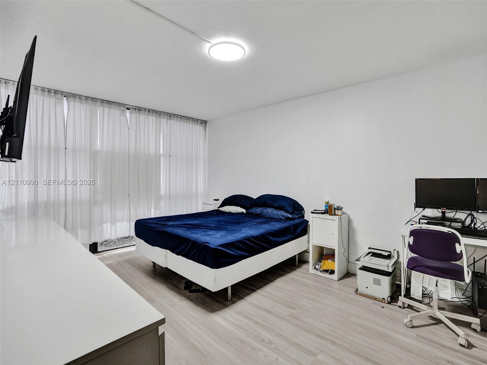 Interior space featuring light-toned wood-finish flooring, floor-to-ceiling window treatments, and a central flush-mount LED light fixture