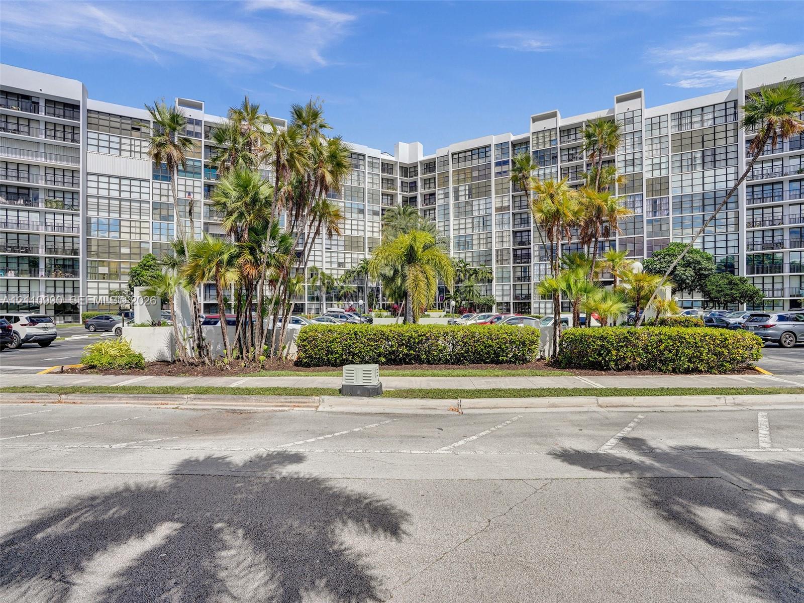 Multi-story concrete residential structure featuring floor-to-ceiling windows, uniform balcony grid, mature palm trees, and manicured shrubbery