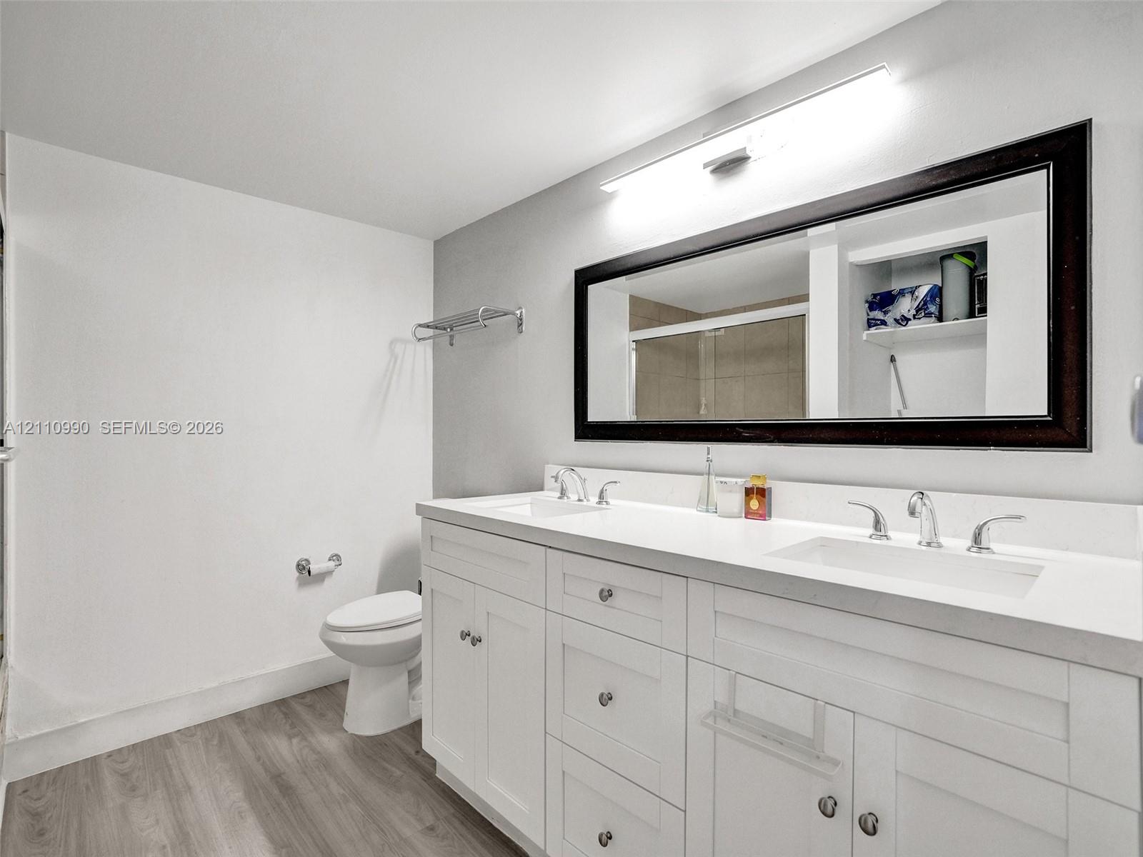 White dual-sink vanity with a solid surface countertop, wood-finish flooring, full-width mirror with a dark frame, and chrome-finish hardware