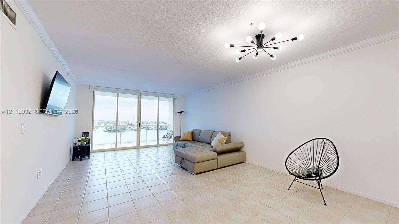 Expansive living area featuring tile flooring, crown molding, a modern sputnik-style chandelier, and floor-to-ceiling sliding glass doors opening to a balcony