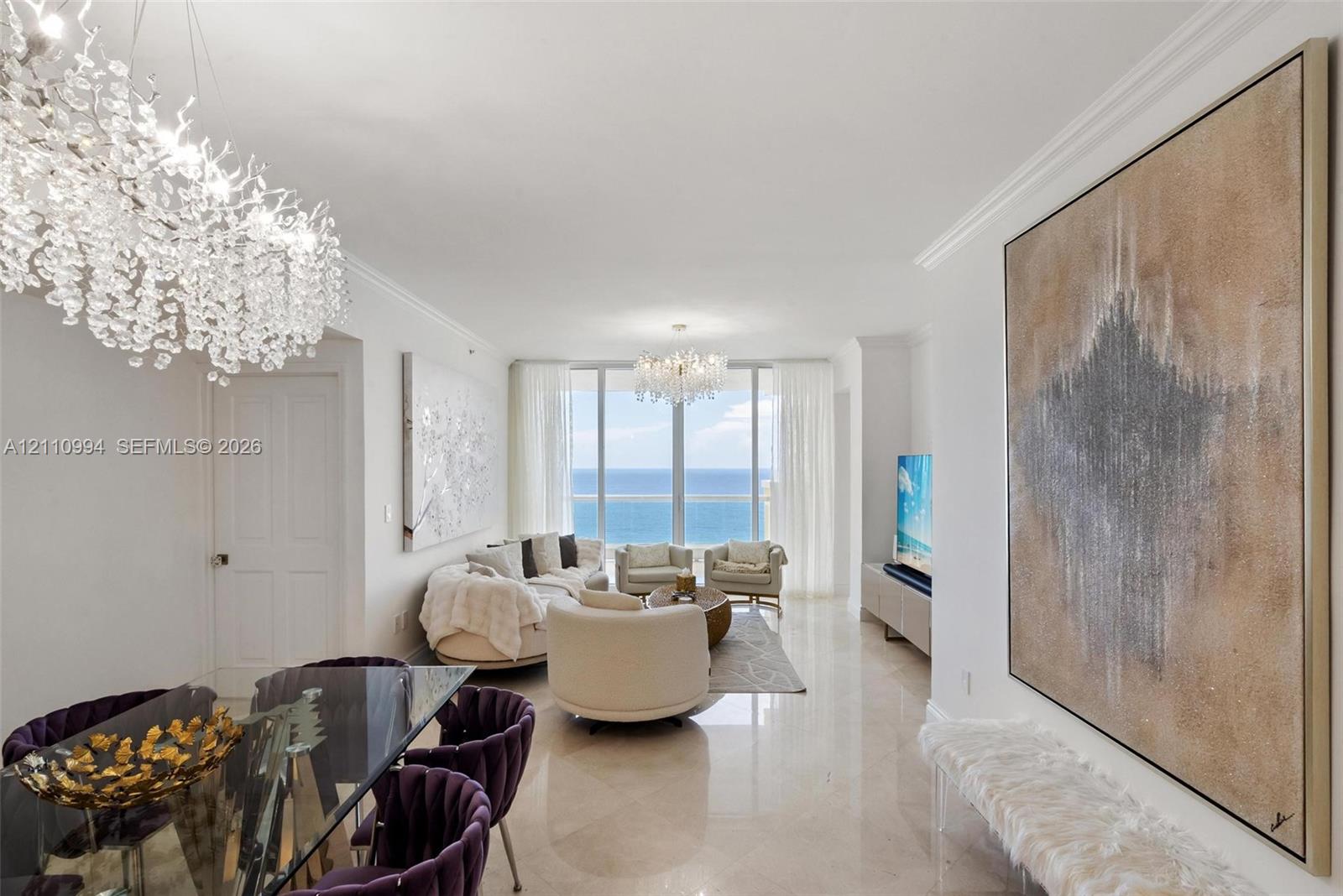 Expansive living area featuring floor-to-ceiling windows with ocean scenery, polished stone flooring, recessed crown molding, and dual crystal chandeliers Expansive living area featuring floor-to-ceiling windows with ocean scenery, polished stone flooring, recessed crown molding, and dual crystal chandeliers