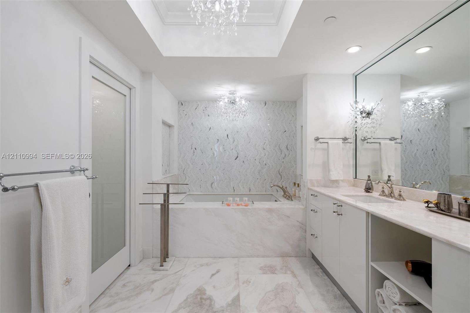 Soaking tub with stone surround, custom tiled accent wall, skylight with recessed tray ceiling, dual crystal chandeliers, and white cabinetry with stone countertop
