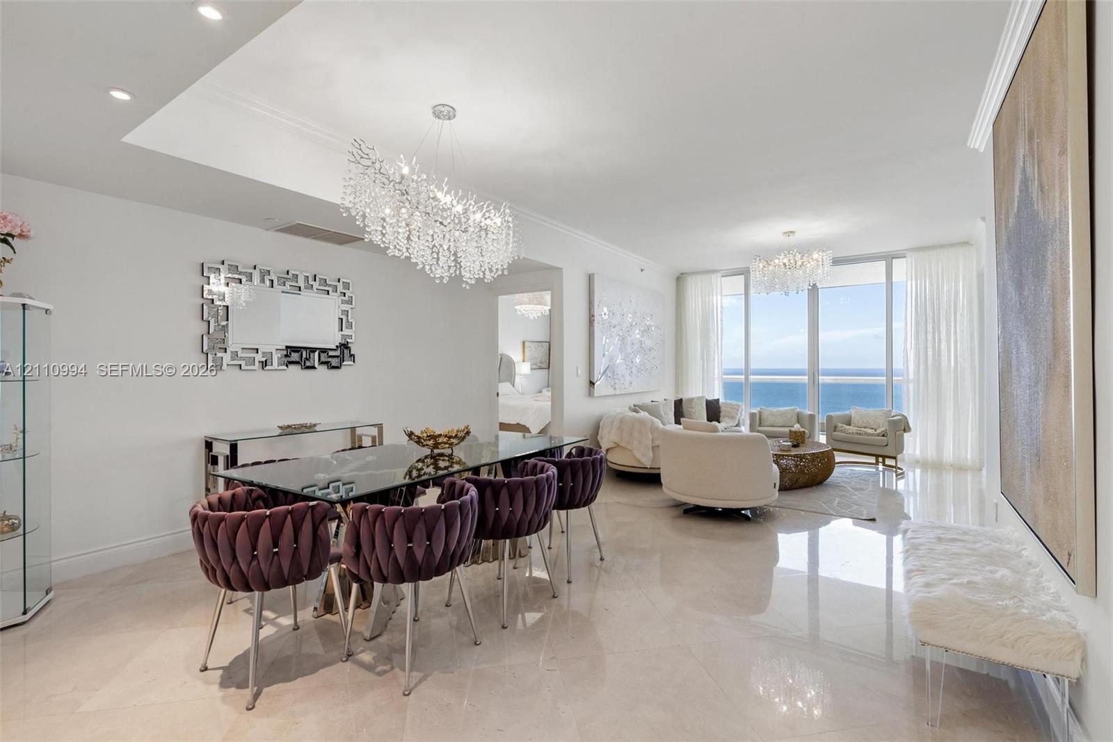 Open-concept interior featuring light-toned polished stone flooring, recessed ceiling lighting, statement crystal chandeliers, and expansive floor-to-ceiling windows with ocean scenery