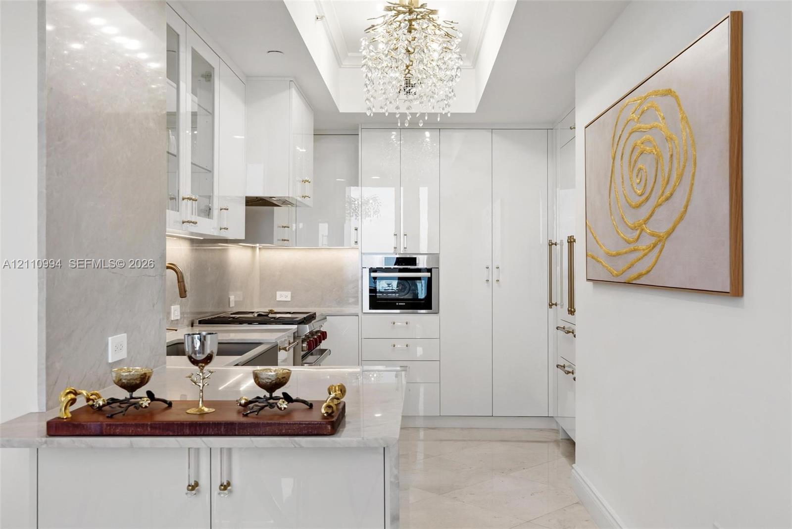 High-gloss white cabinetry featuring polished hardware, stone-finish countertops, recessed tray ceiling with crystal-style chandelier, and built-in stainless steel wall oven