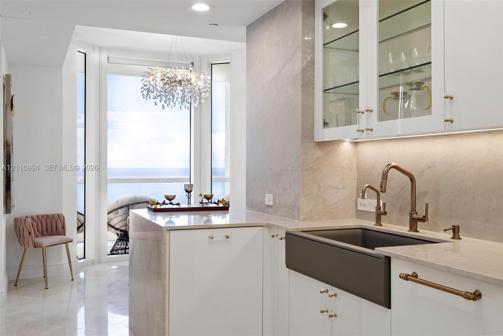 Custom kitchen featuring white cabinetry with glass-front uppers, stone-surround backsplash, farmhouse sink with bronze-tone hardware, and light-reflecting flooring