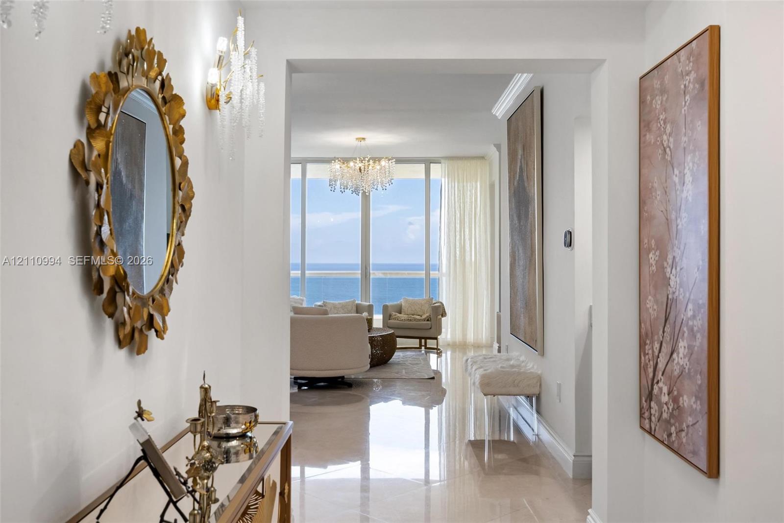 Entryway featuring polished stone-finish flooring, crown molding, and crystal wall-mounted light fixtures, leading to floor-to-ceiling windows with ocean scenery