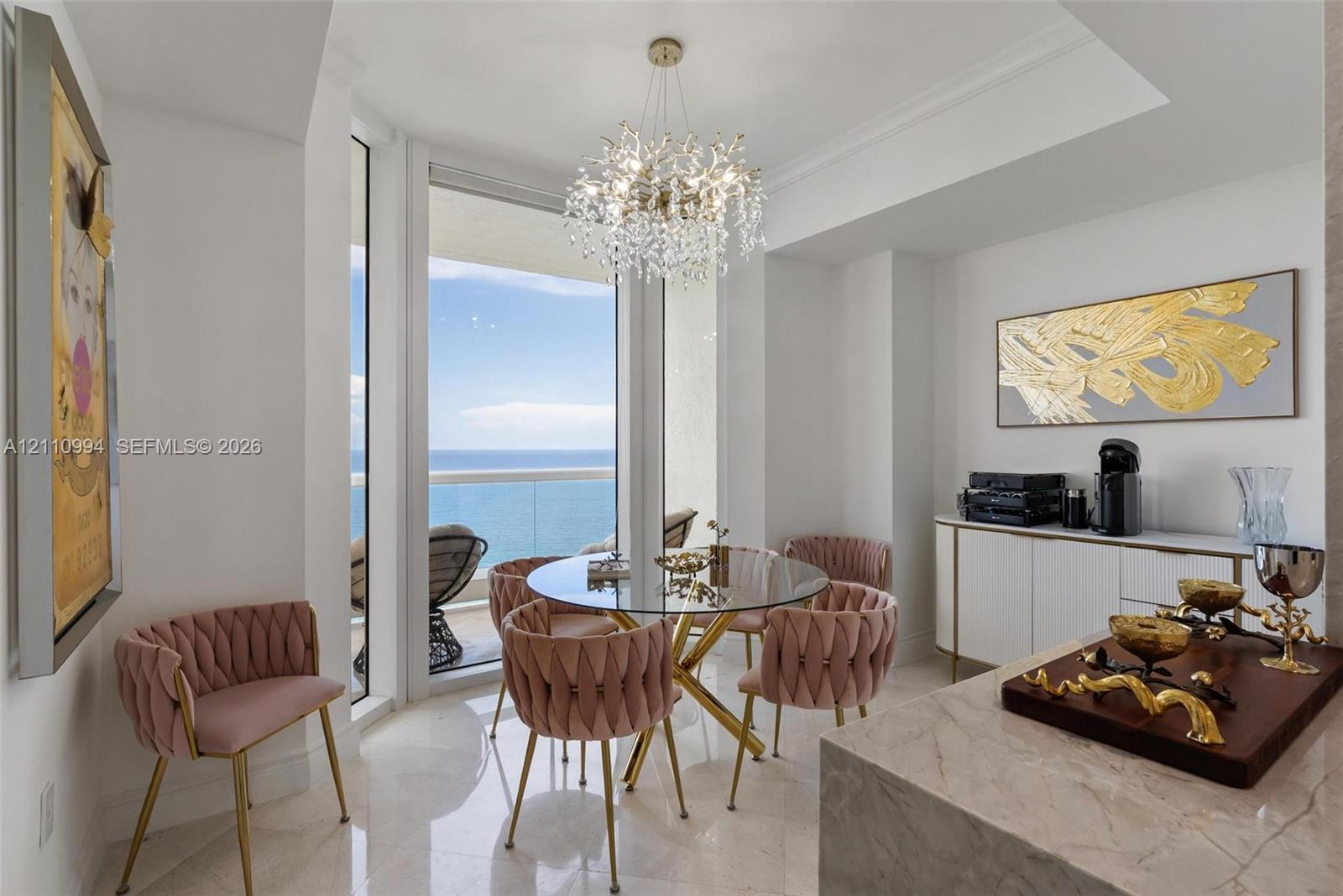 Dining area featuring large-format polished stone flooring, floor-to-ceiling windows with ocean horizon, a multi-tiered crystal chandelier, and built-in white cabinetry with gold-tone hardware Dining area featuring large-format polished stone flooring, floor-to-ceiling windows with ocean horizon, a multi-tiered crystal chandelier, and built-in white cabinetry with gold-tone hardware