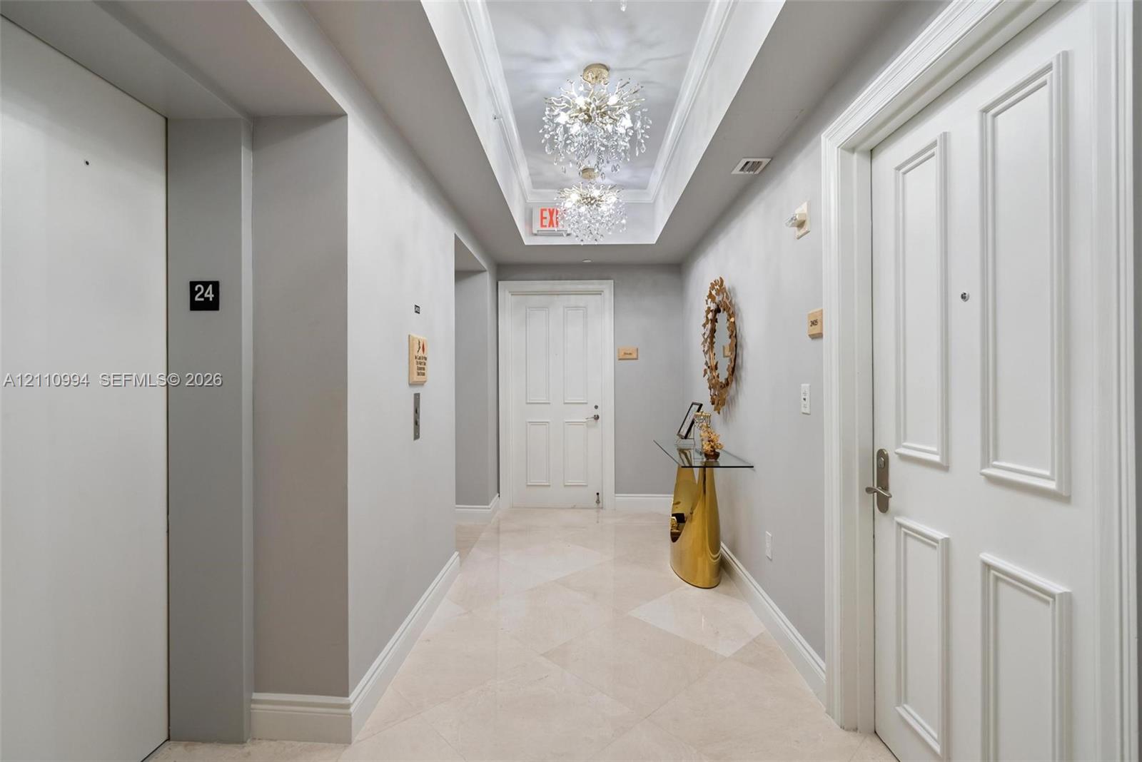 Hallway featuring stone tile flooring, crown molding, dual crystal chandelier light fixtures set within a tray ceiling, and panel-style entry doors