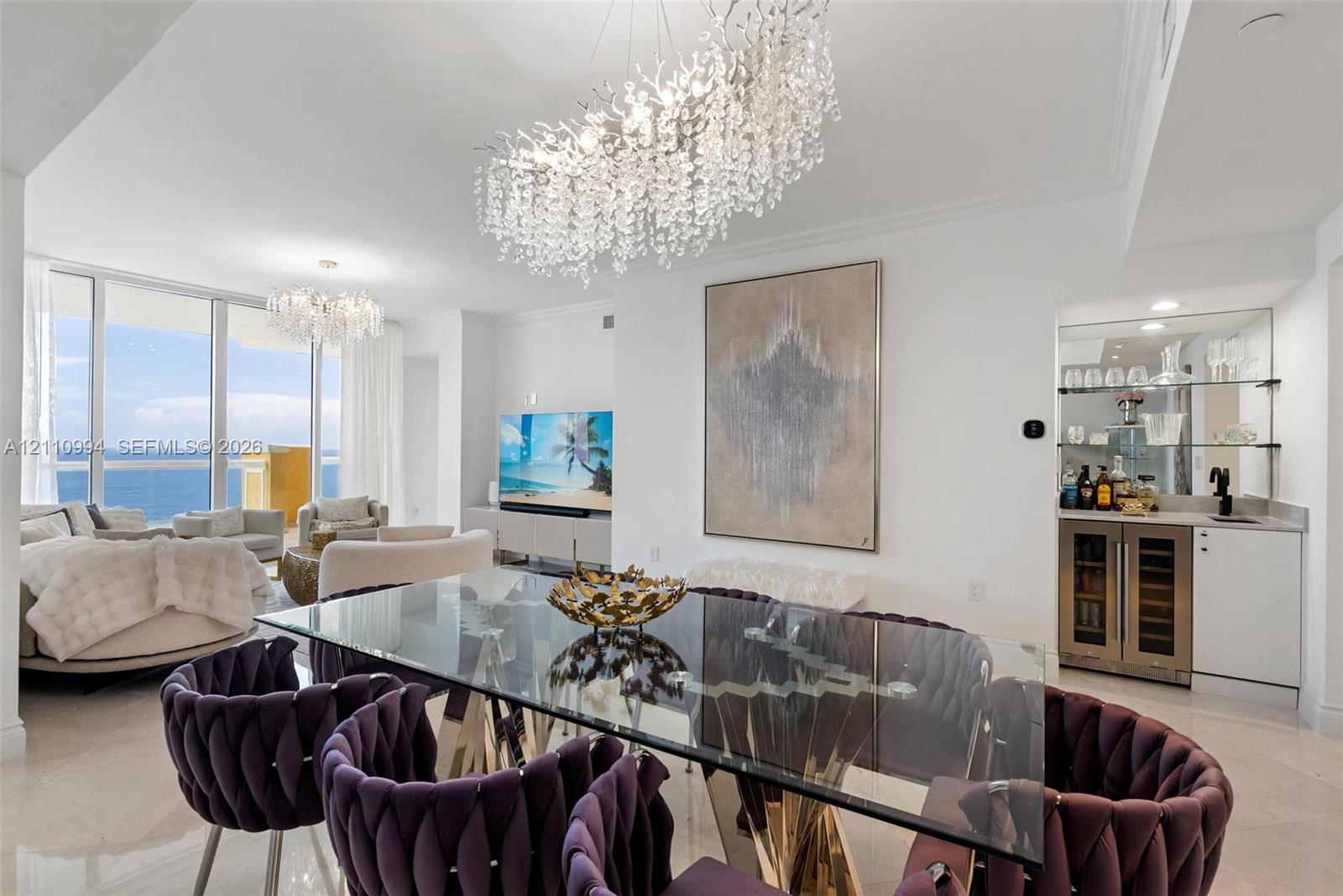 Open-concept space featuring floor-to-ceiling glass, custom crystal chandeliers, polished stone flooring, and a built-in wet bar with a glass-front beverage cooler