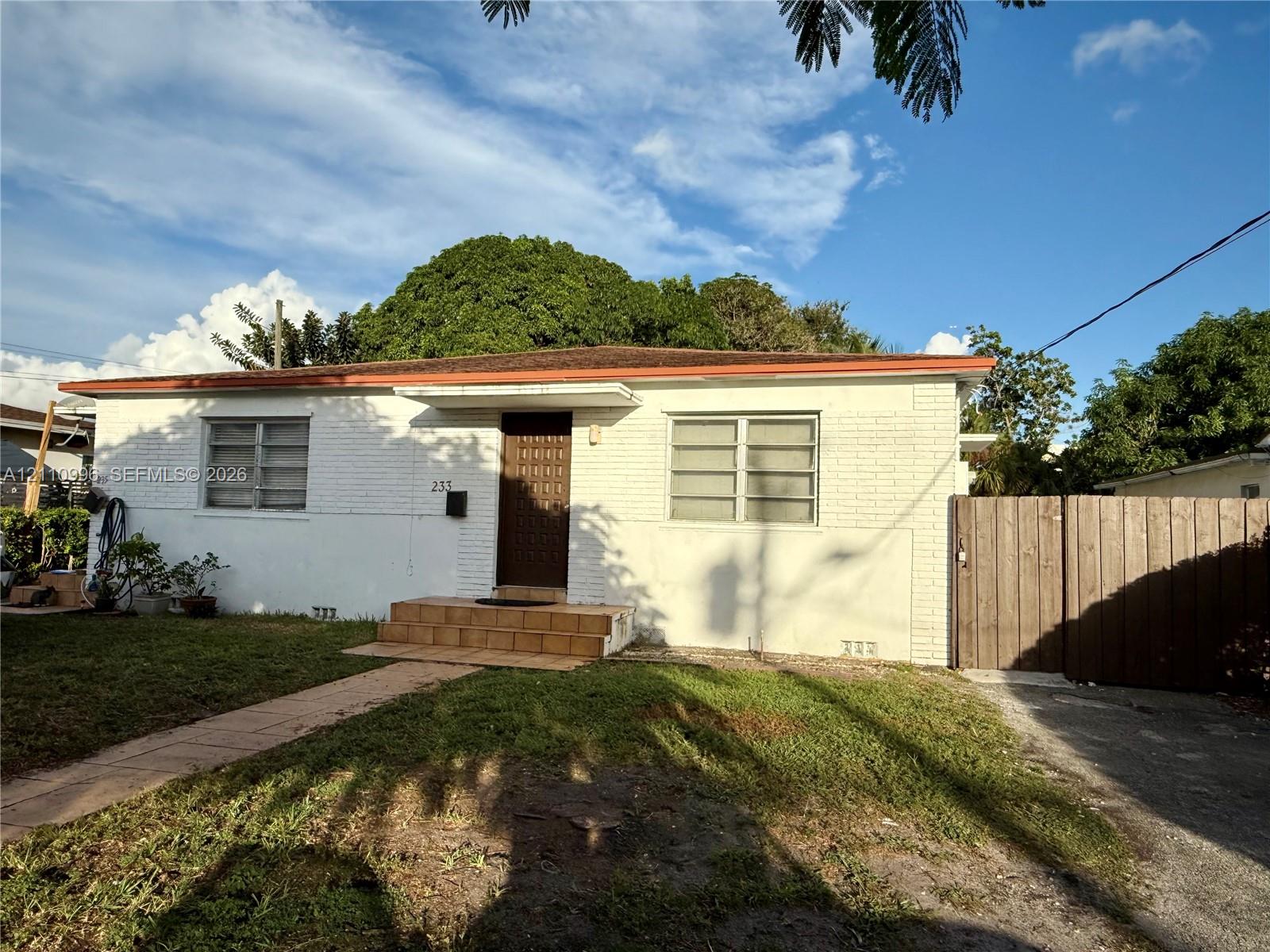 Single-story residence featuring white painted brick exterior, horizontal sliding windows, a concrete walkway, and a full-height wooden privacy fence Single-story residence featuring white painted brick exterior, horizontal sliding windows, a concrete walkway, and a full-height wooden privacy fence