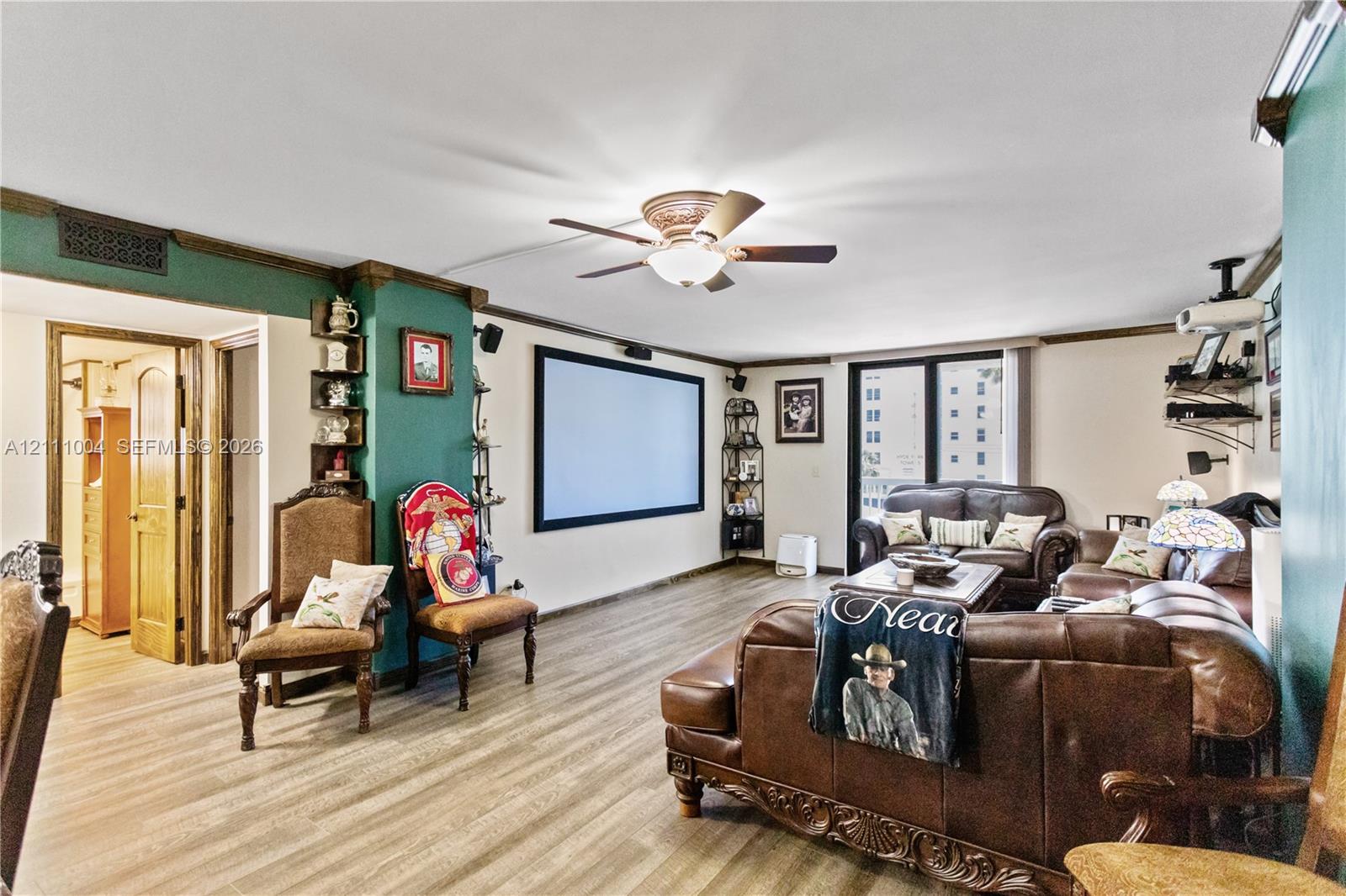 Living area featuring wood-finish flooring, a ceiling fan with integrated lighting, built-in corner shelving, and a wall-mounted projection screen