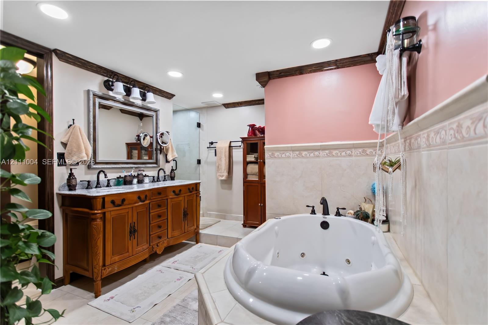 Bathroom featuring a custom wood vanity with a dual-sink stone countertop, a jetted soaking tub, tiled wainscoting, and recessed lighting