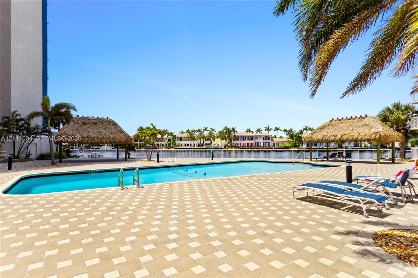Swimming pool area featuring a paved deck, dual thatch-roofed cabanas, and perimeter fencing