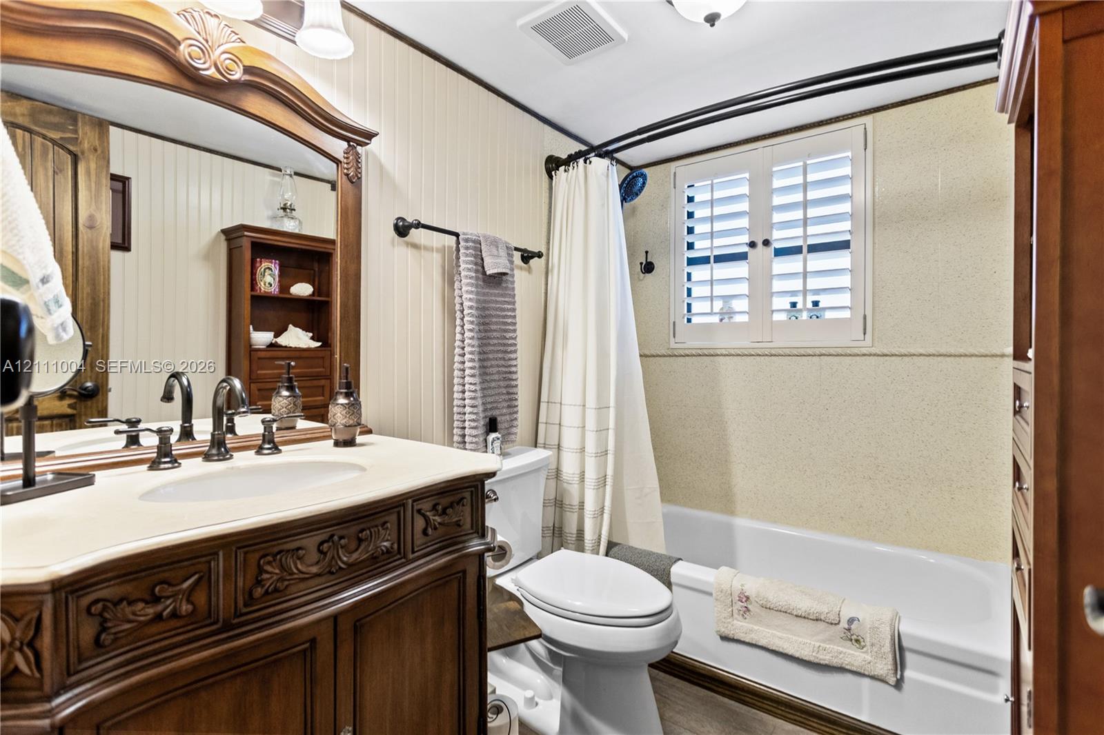 Bathroom featuring a dark wood-finish vanity with an integrated basin, beadboard wall paneling, bathtub with white shutter-style window treatments, and a decorative arched mirror frame