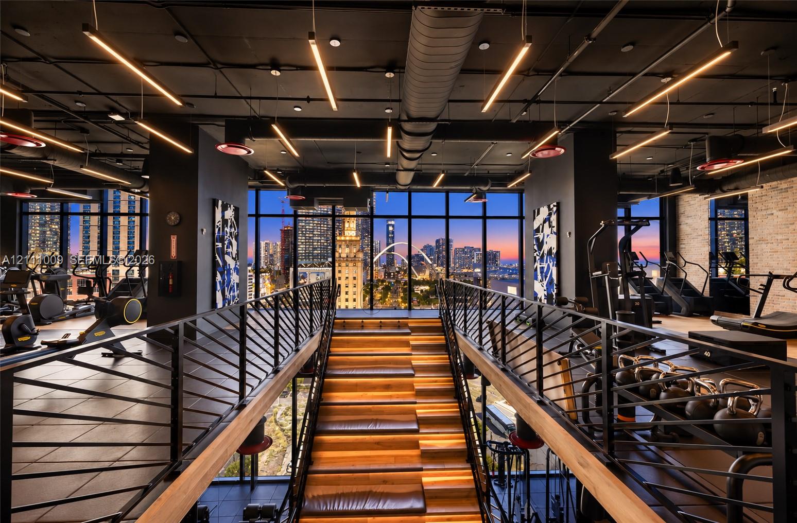 Industrial-style space featuring floor-to-ceiling windows, exposed ductwork, metal railings, wood-tread staircase with integrated lighting, and brick accent wall