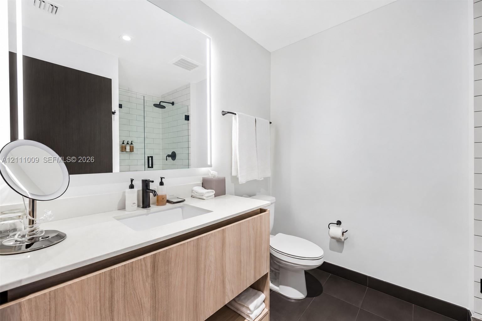 Floating vanity with wood-finish cabinetry, white countertop, integrated rectangular sink, matte black faucet, and large LED-backlit mirror