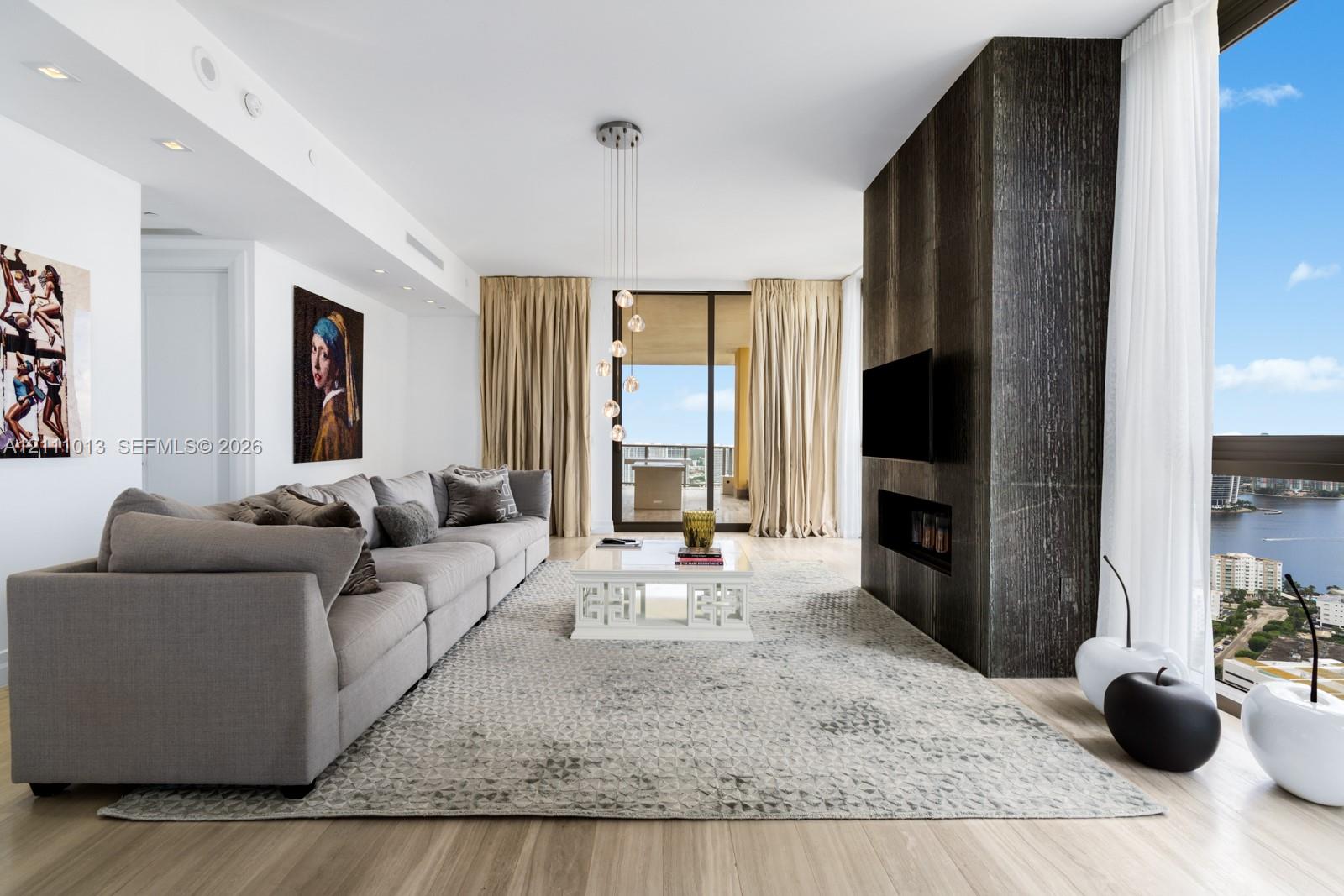 Floor-to-ceiling windows, dark stone-finish accent wall with integrated fireplace, light-toned wood-finish flooring, recessed ceiling lighting, and multi-pendant light fixture