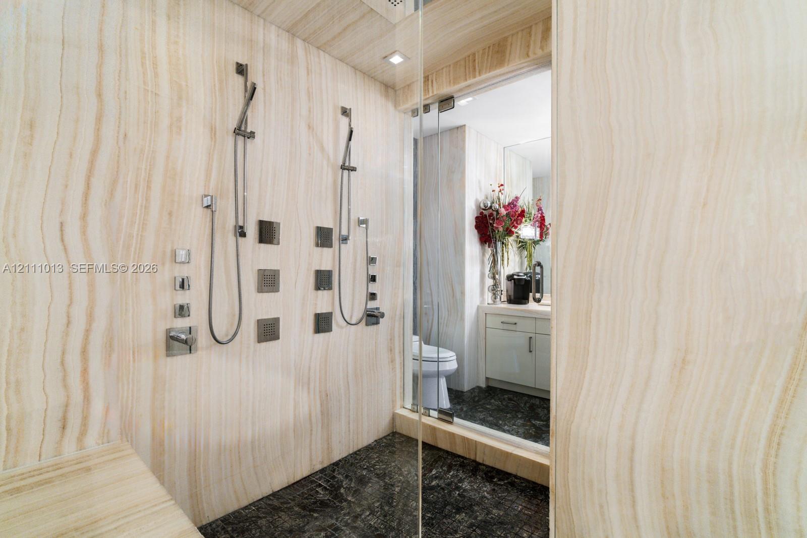 Walk-in shower featuring full-height natural stone wall cladding, dual handheld shower heads with integrated body sprayers, a built-in stone bench, and glass enclosure