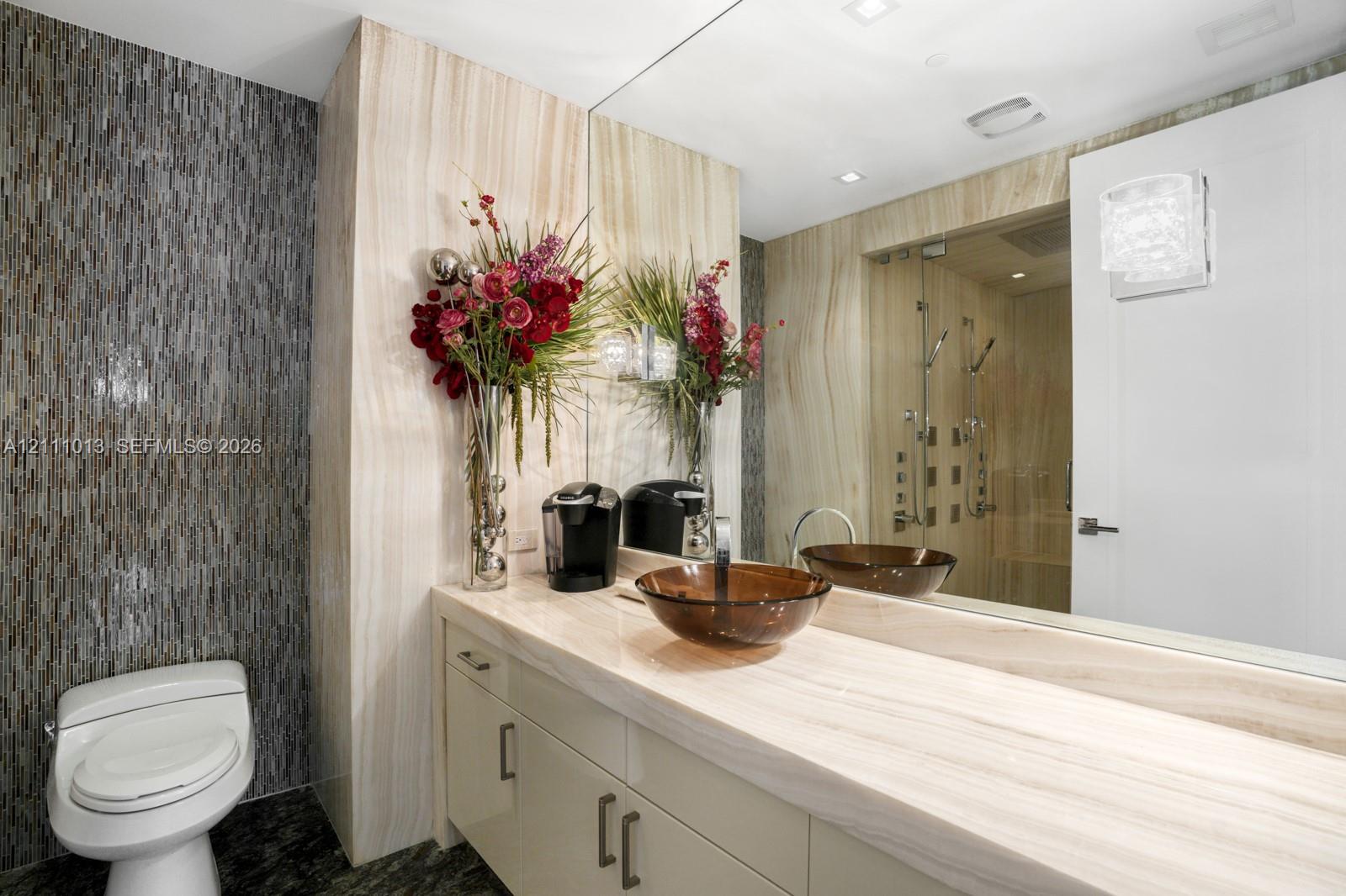 Bathroom featuring a stone slab countertop, glass vessel sink, full-width mirror, and a glass-enclosed shower with a multi-head fixture