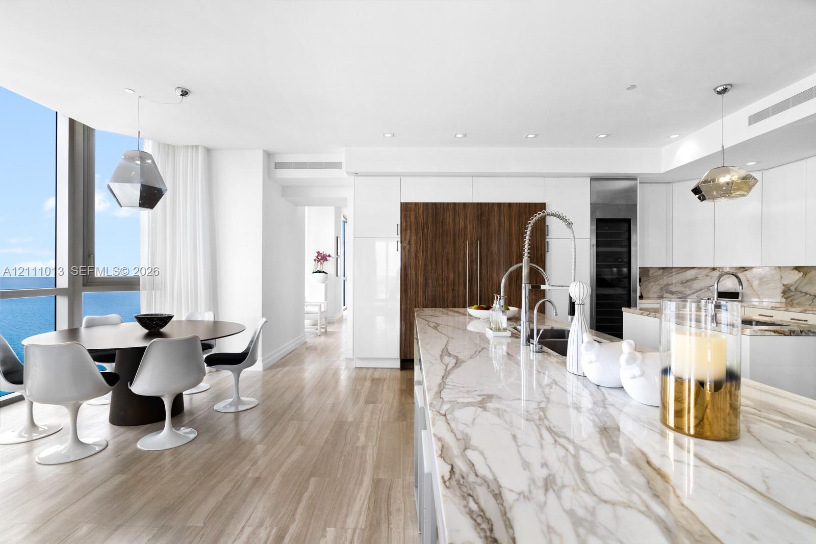 Contemporary kitchen featuring a large stone island with an integrated sink, high-gloss cabinetry, wood-finish flooring, and floor-to-ceiling windows