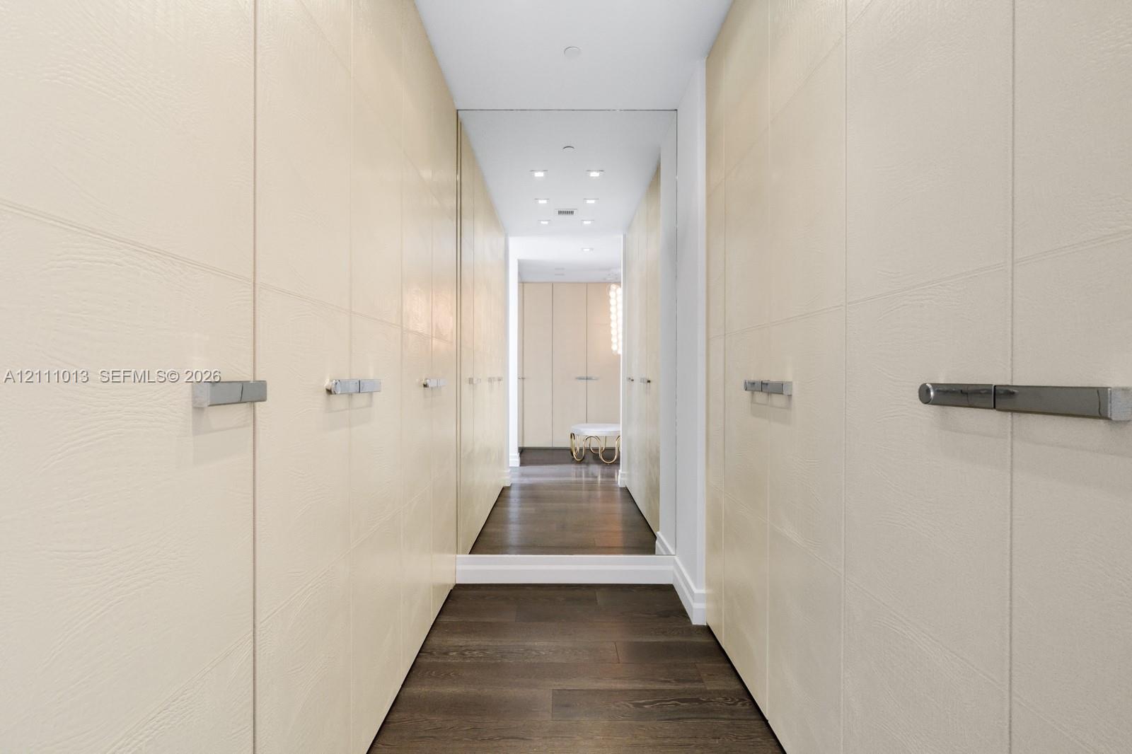 Custom floor-to-ceiling cabinetry with textured cream-colored finish, polished metal hardware, full-height wall mirrors, recessed ceiling lighting, and dark wood-finish flooring