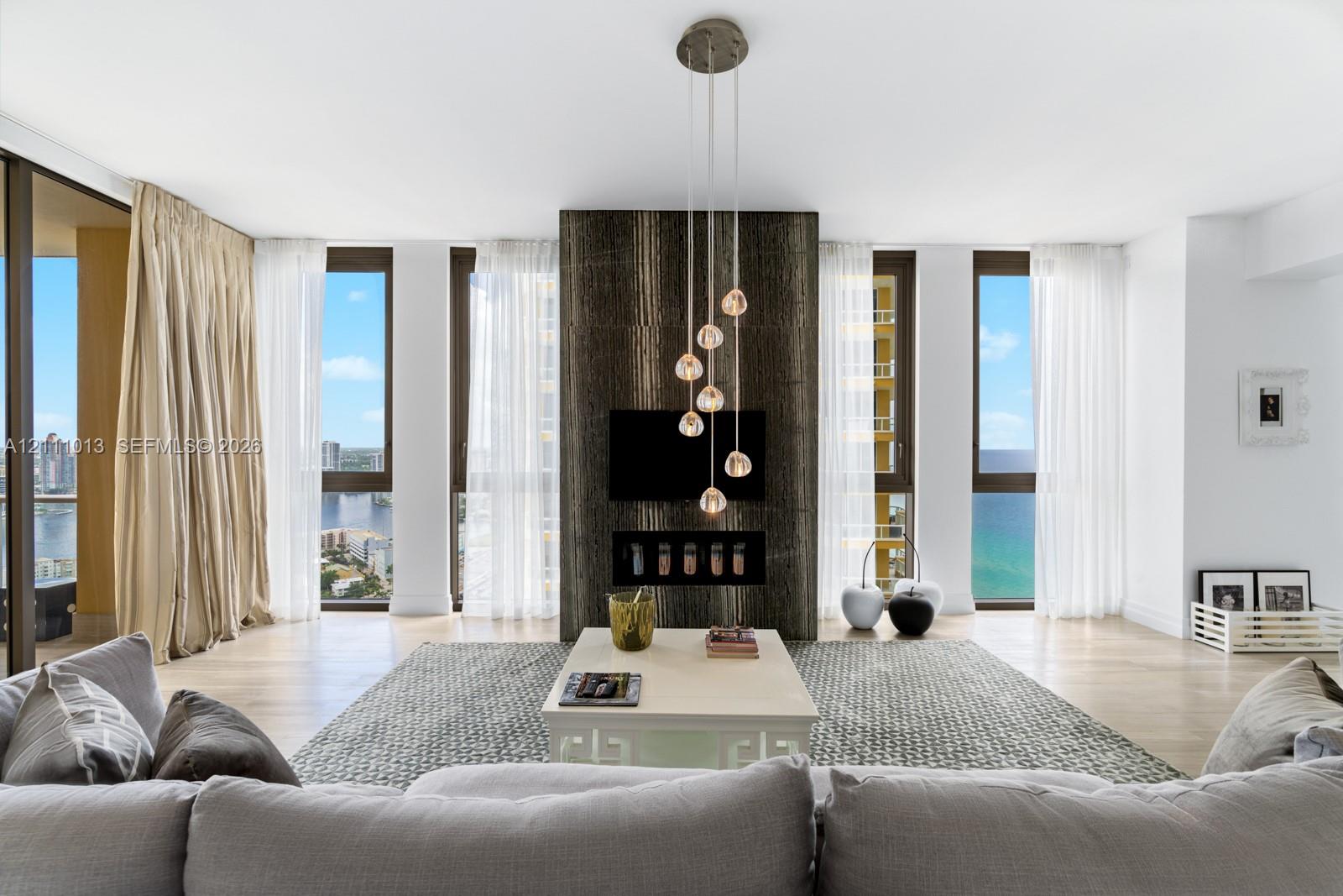 Floor-to-ceiling windows, stone-clad fireplace feature wall, wood-finish flooring, and multi-pendant lighting fixture