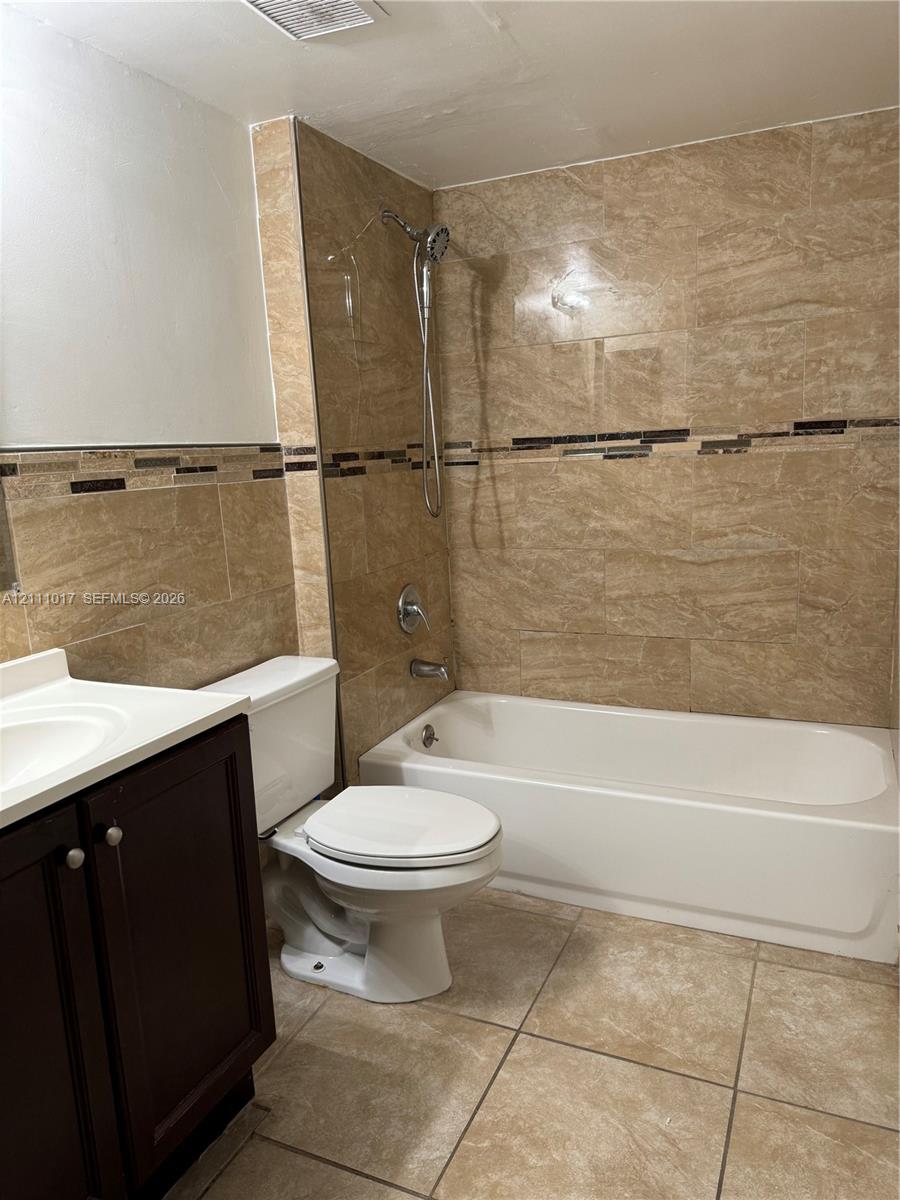 Bathroom featuring floor-to-ceiling tile surround with decorative glass accent trim, integrated tub and shower combination, dark-finish cabinetry, and neutral tile flooring