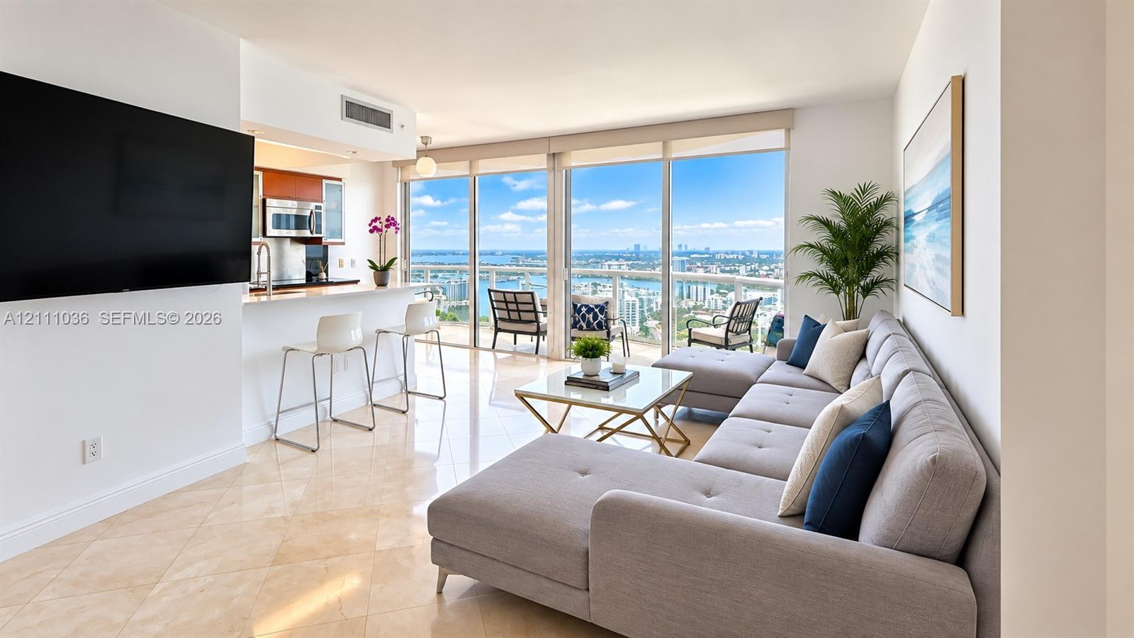 Open-concept interior featuring large-format tile flooring, floor-to-ceiling windows, an integrated kitchen breakfast bar, and a balcony with expansive urban and water scenery