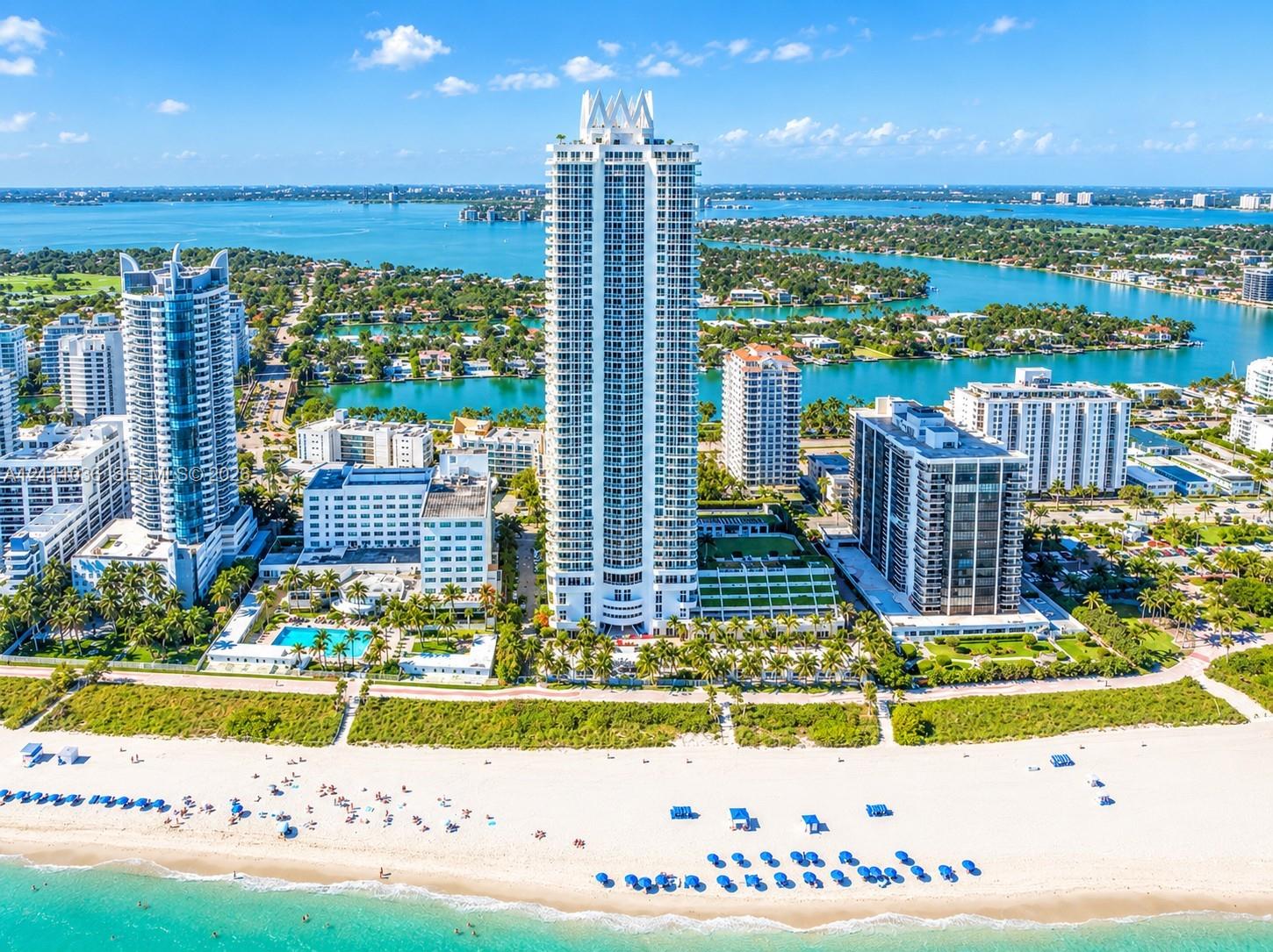 High-rise residential tower featuring a white concrete exterior, tiered architectural design, expansive glass balcony railings, direct sand beach access, and surrounding coastal waterfront scenery