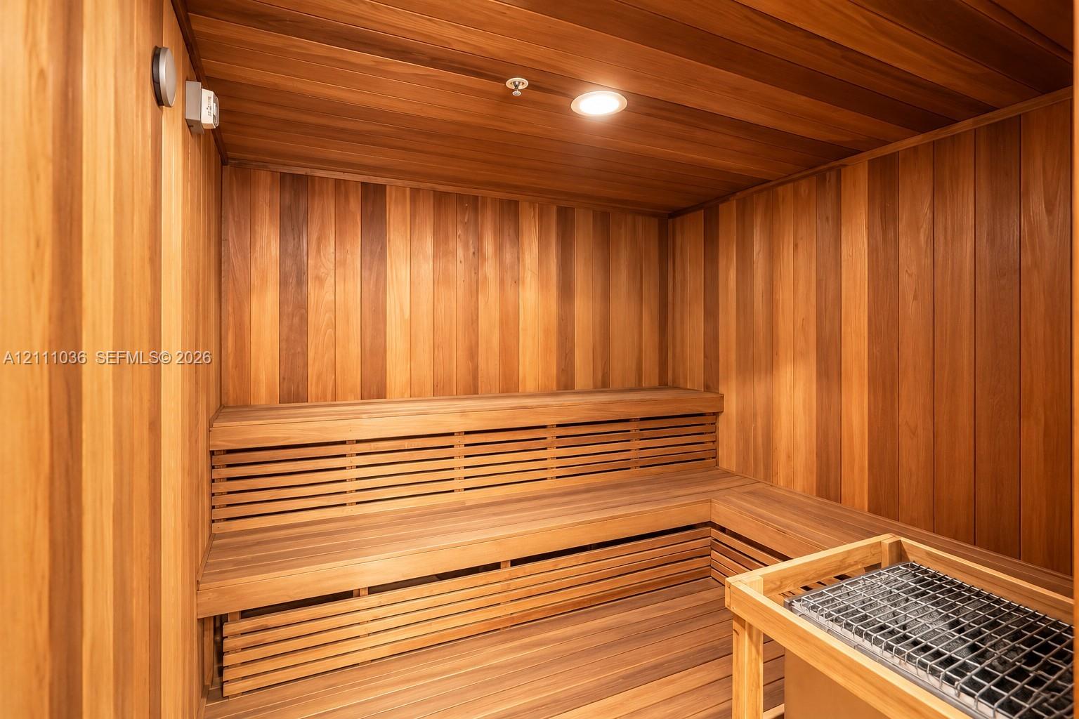 Sauna featuring tongue-and-groove wood paneling, multi-level built-in bench seating, recessed ceiling lighting, and a stone-filled heating unit with a wooden safety enclosure
