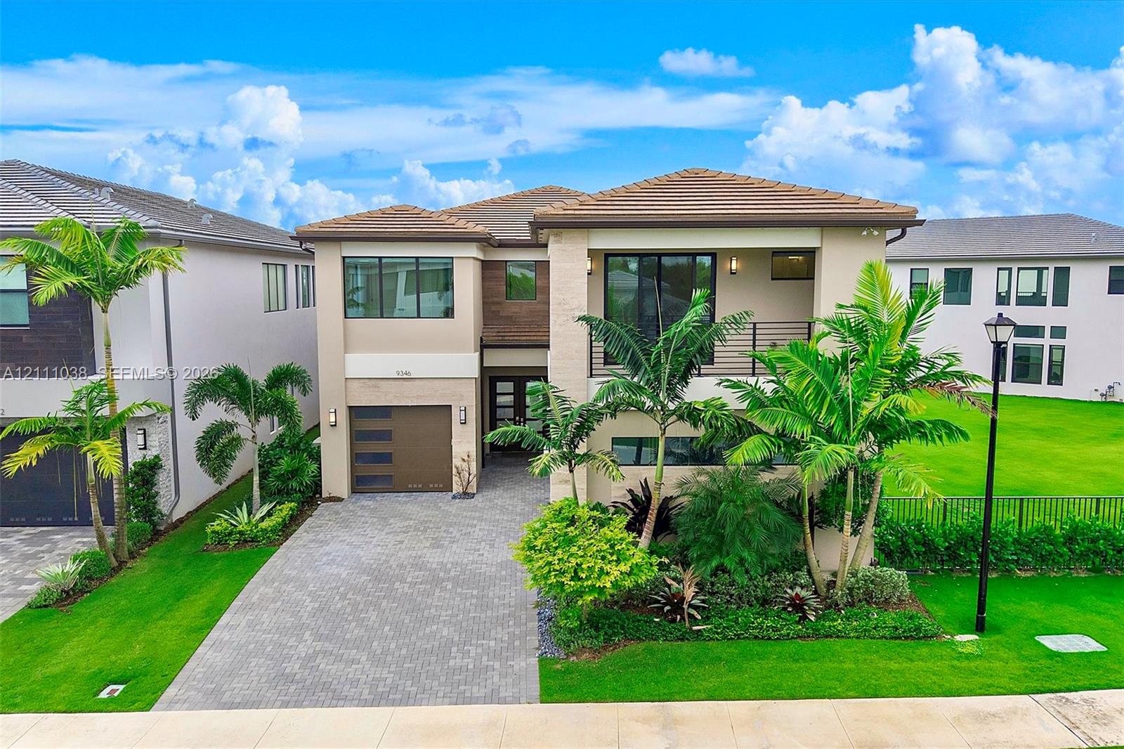 Two-story residence featuring a tile roof, paver driveway, integrated garage door with horizontal glass panels, upper-level balcony with metal railing, and tropical landscaping