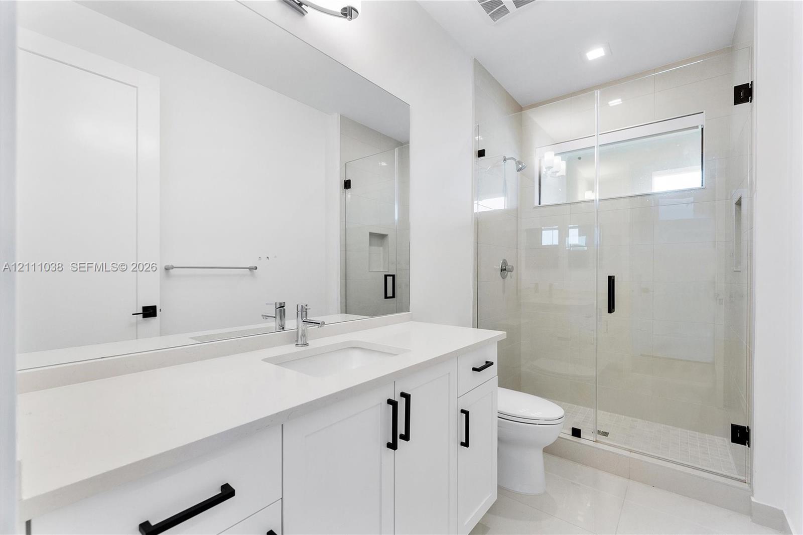 White cabinetry vanity with solid surface countertop, glass-enclosed shower with tiled walls and built-in niche, and matte black hardware fixtures