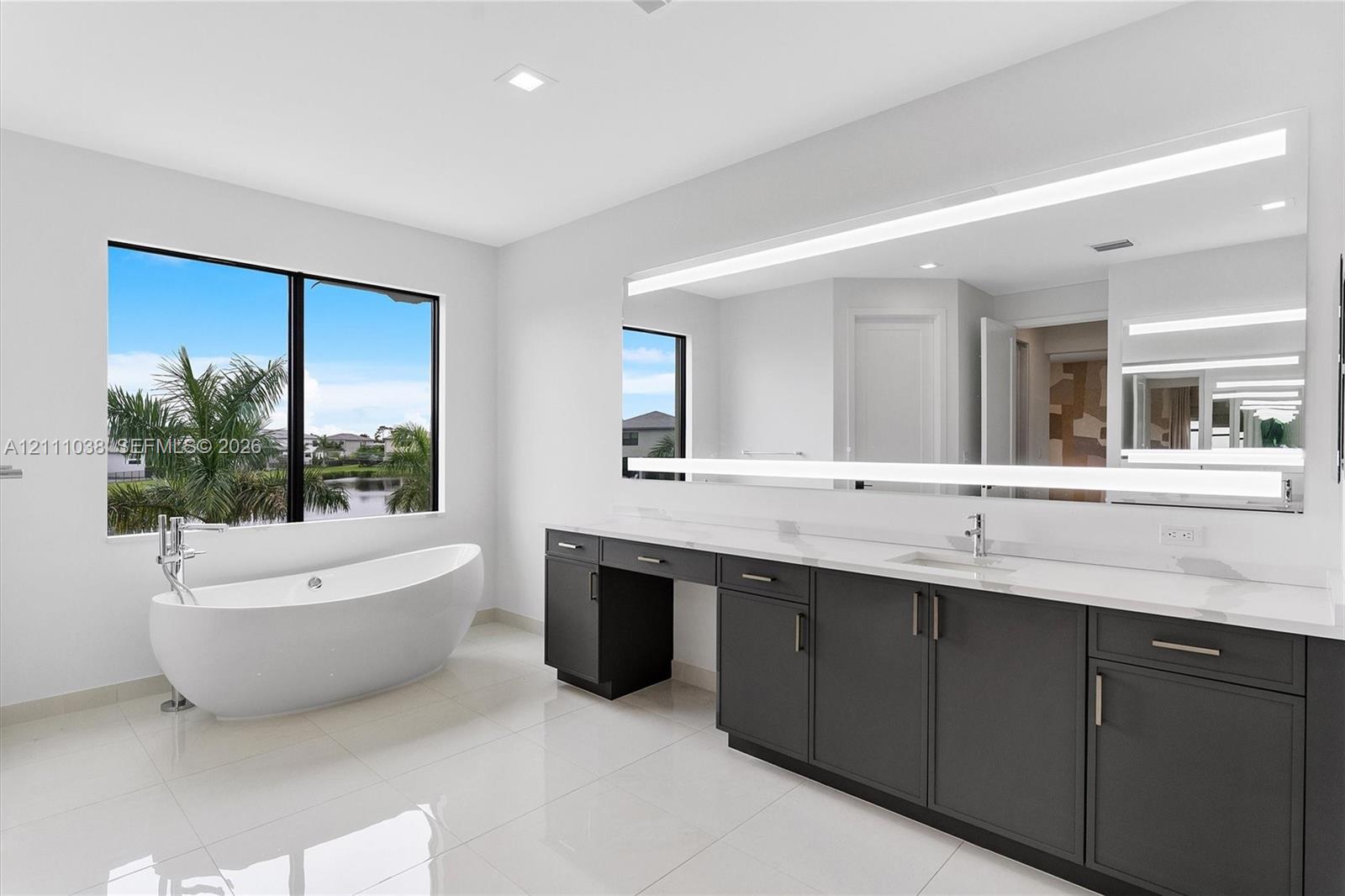 Freestanding soaking tub with floor-mounted faucet, dual-sink vanity with light-tone countertops, dark cabinetry, and oversized LED-backlit mirror