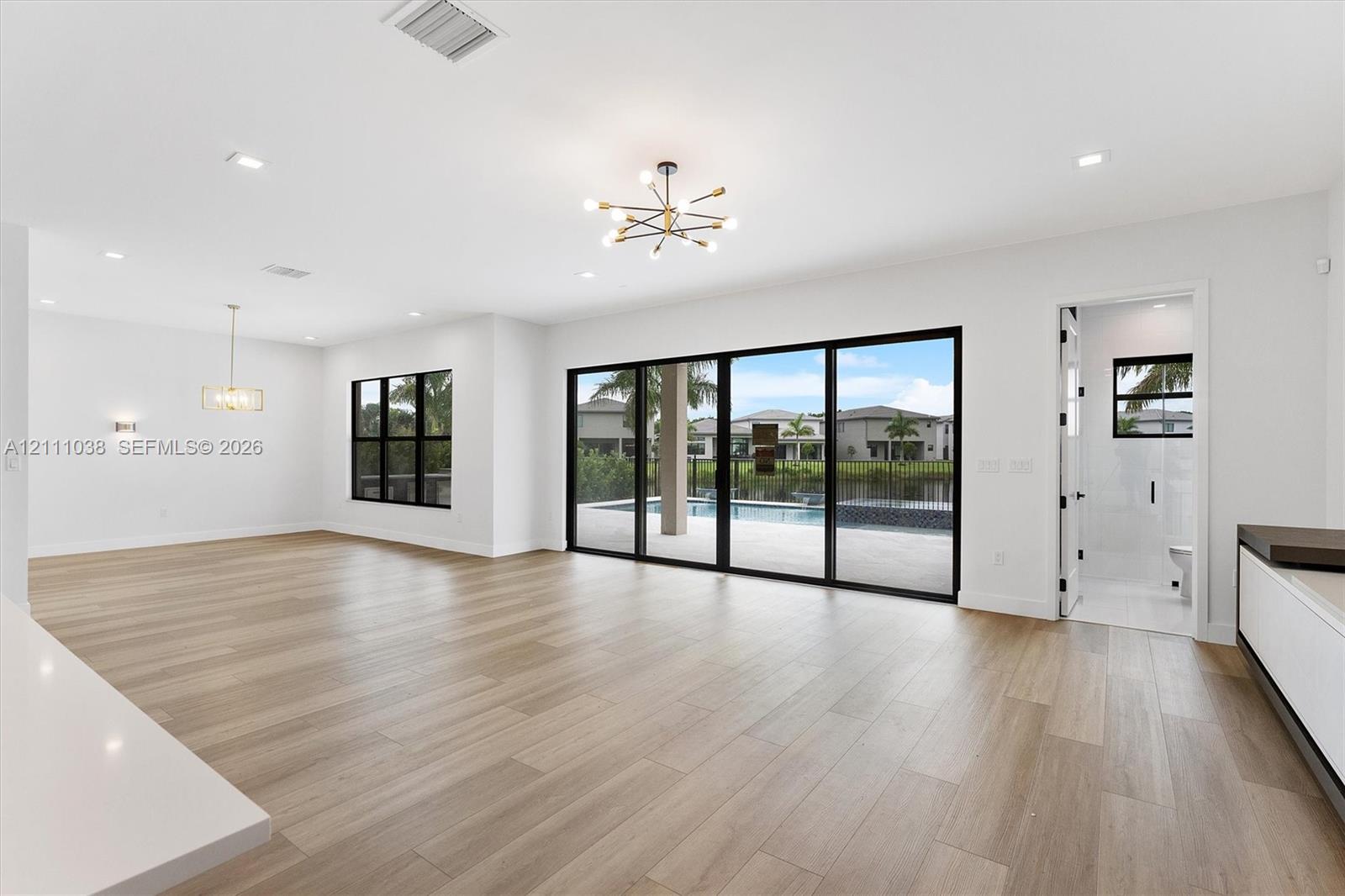 Open-concept interior featuring light wood-finish flooring, multi-panel sliding glass doors with black frames, recessed lighting, and modern multi-arm chandeliers