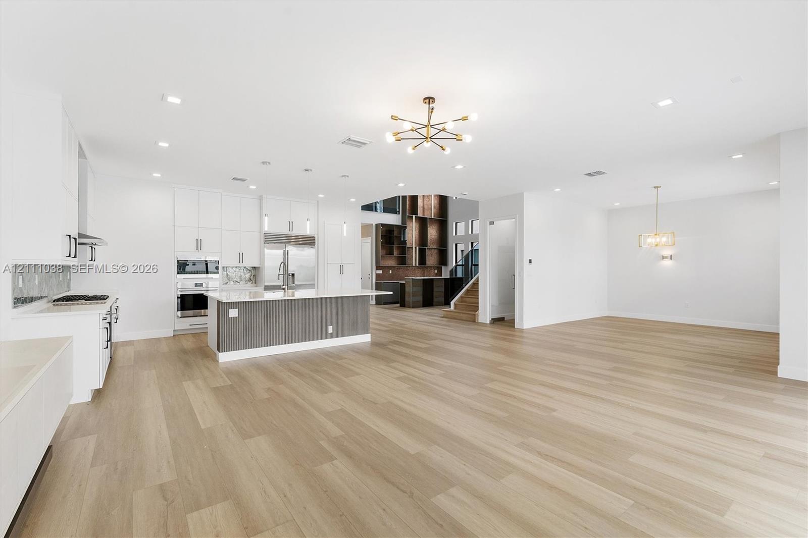 Open-concept area featuring wood-finish flooring, a kitchen island with stone countertop, white cabinetry with contrast base, modern chandelier lighting, and recessed ceiling lights