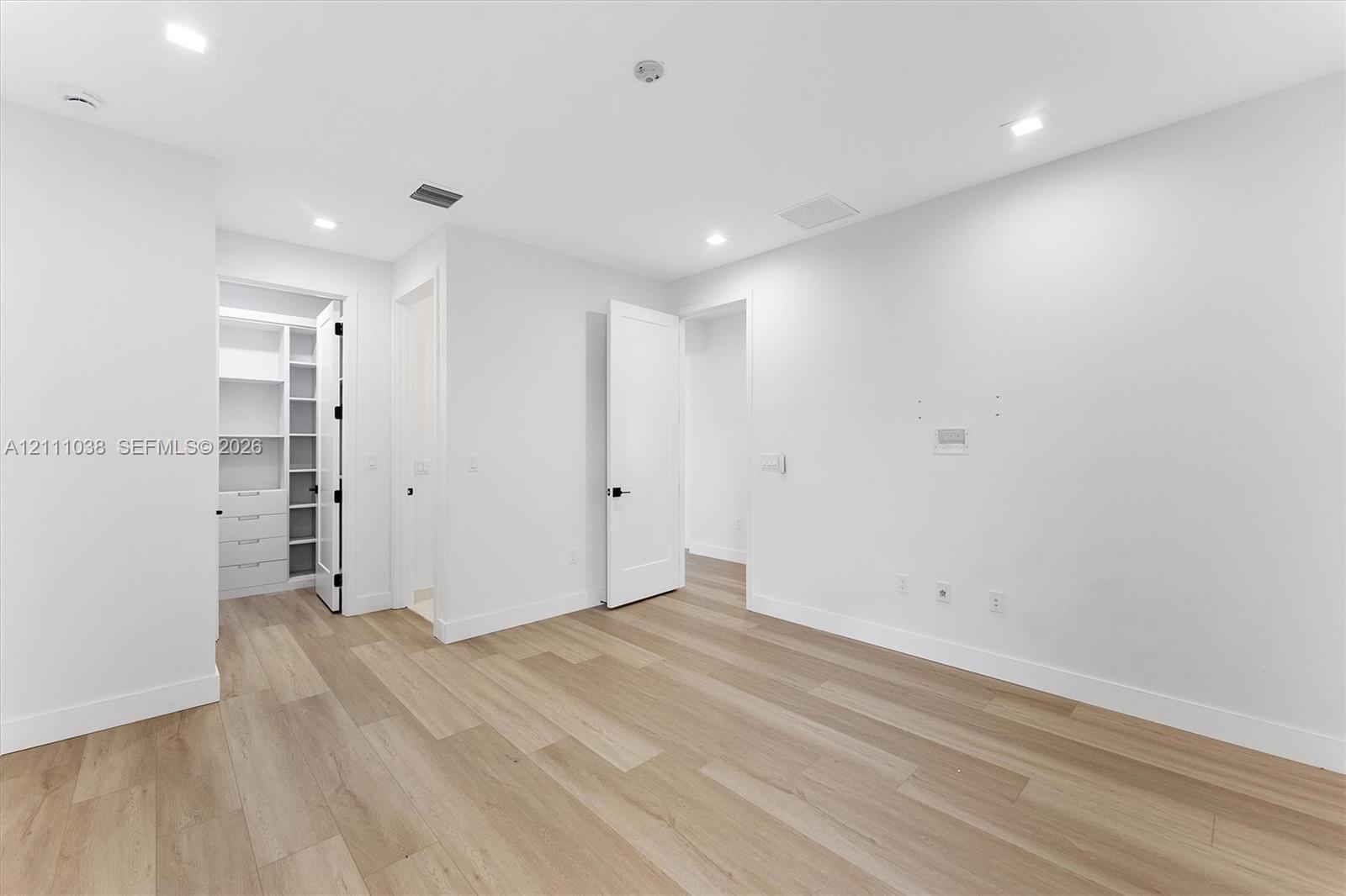 Neutral-toned space featuring wood-finish flooring, recessed lighting, shaker-style doors with matte black hardware, and an integrated walk-in closet with built-in shelving