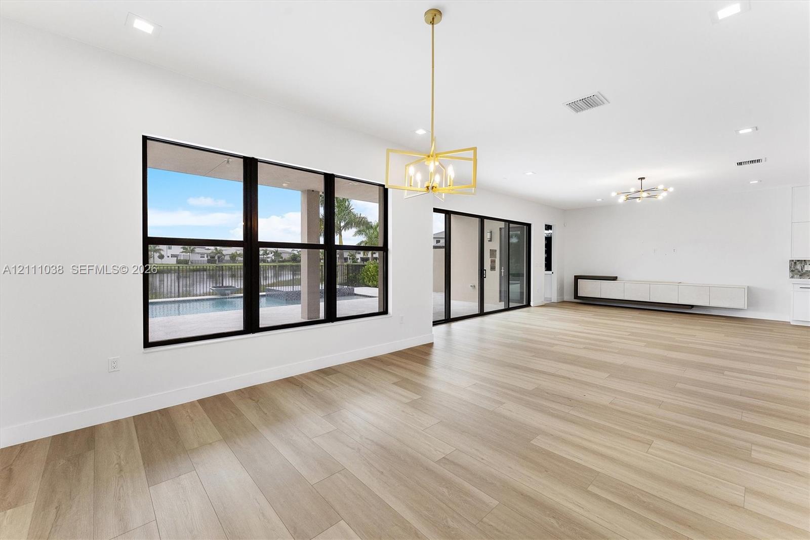 Open-concept interior featuring light wood-finish flooring, large multi-pane windows with dark frames, a geometric metallic pendant light, recessed ceiling lighting, and a built-in wall-mounted cabinet