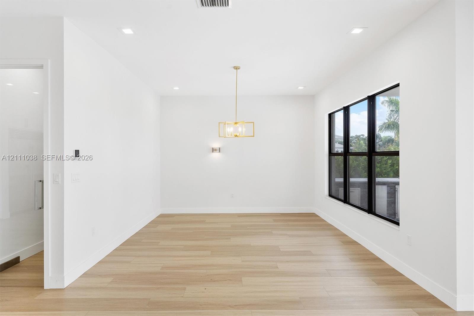 Open-concept interior featuring light wood-finish flooring, recessed lighting, a modern gold-tone geometric chandelier, and floor-to-ceiling windows with black framing