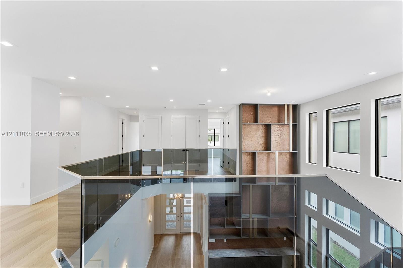 Open landing featuring wood-finish flooring, recessed lighting, floor-to-ceiling glass panel railing, and built-in vertical shelving with textured metallic paneling