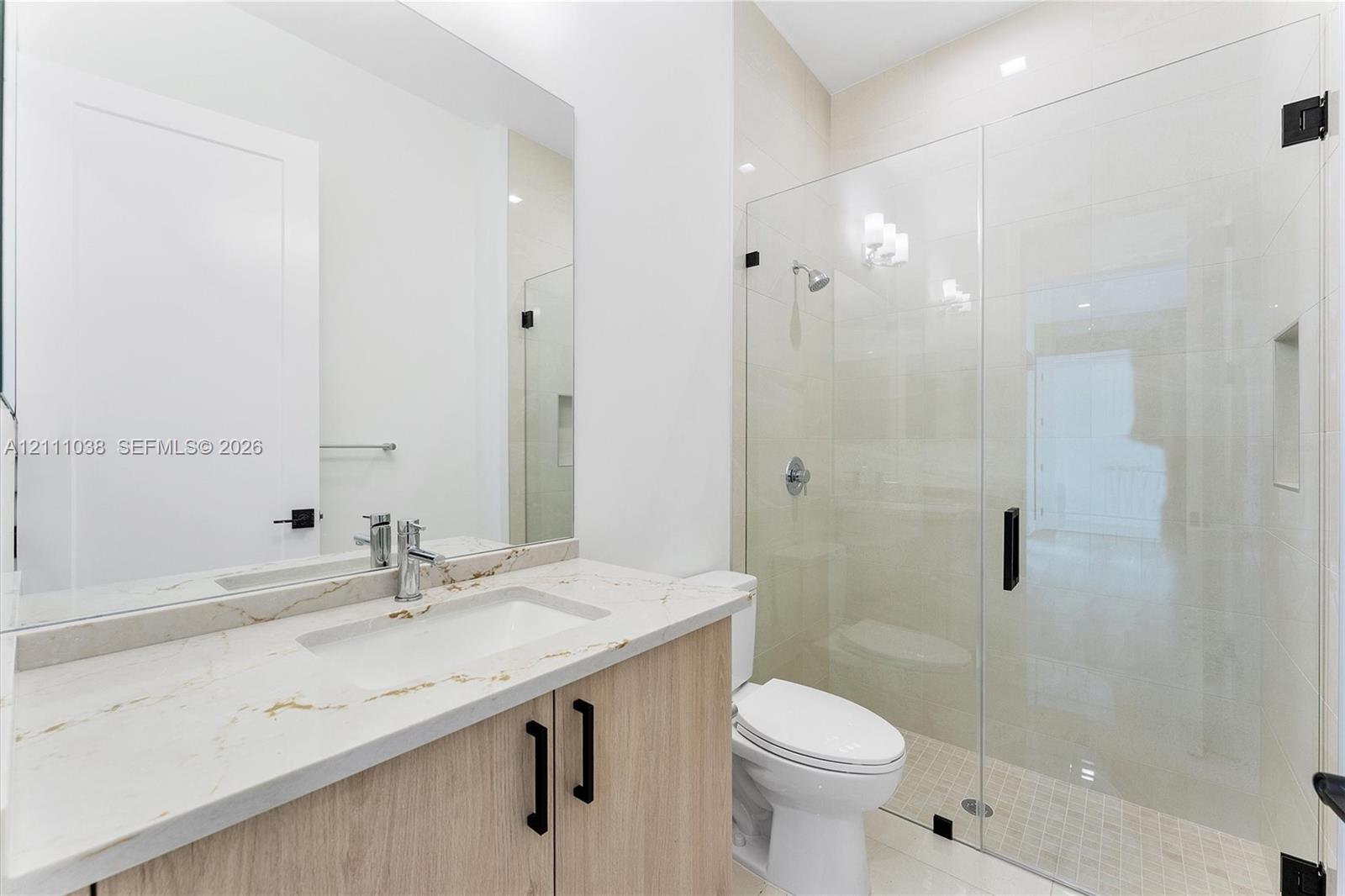 Bathroom featuring a vanity with light wood-finish cabinetry, a stone-surface countertop with an integrated rectangular sink, a large wall-mounted mirror, and a glass-enclosed shower with a recessed wall niche