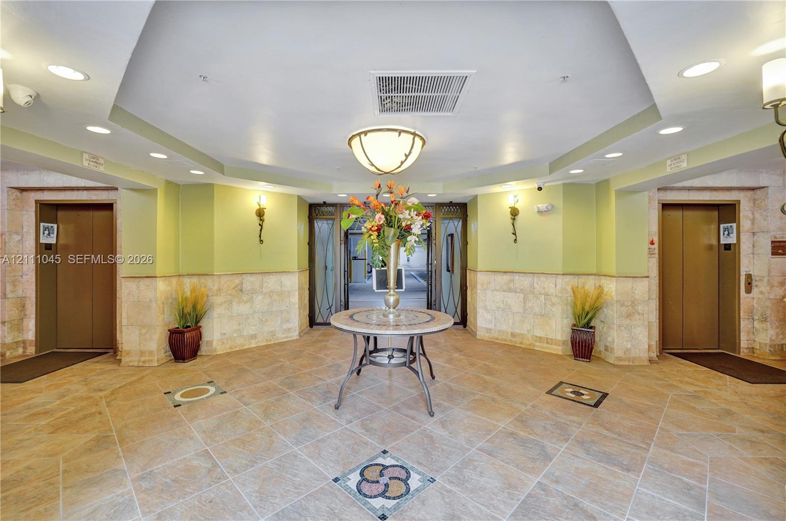 Lobby featuring octagonal tray ceiling with recessed lighting, stone tile flooring with decorative inlay, dual elevators with metallic finish doors, and lower-wall stone masonry cladding