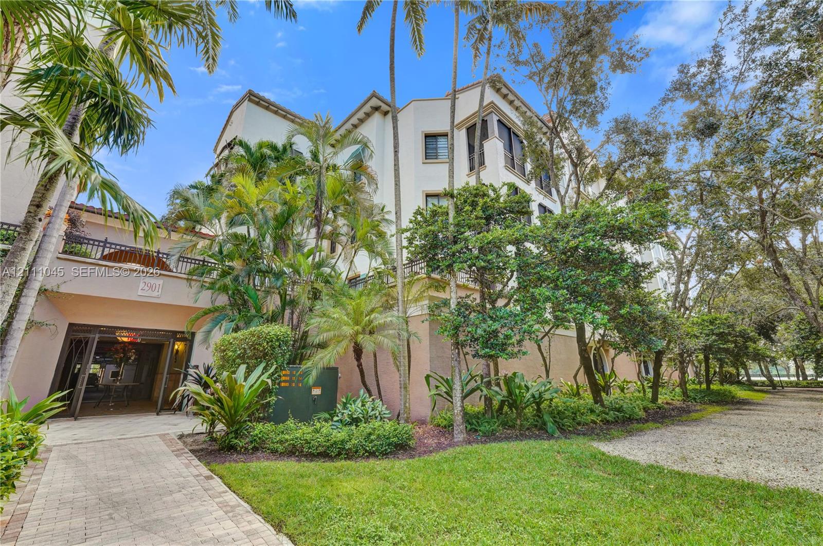 Multi-story exterior featuring light-toned stucco facade, decorative upper-level balconies, paver stone walkway, and established landscaping with mature palm trees
