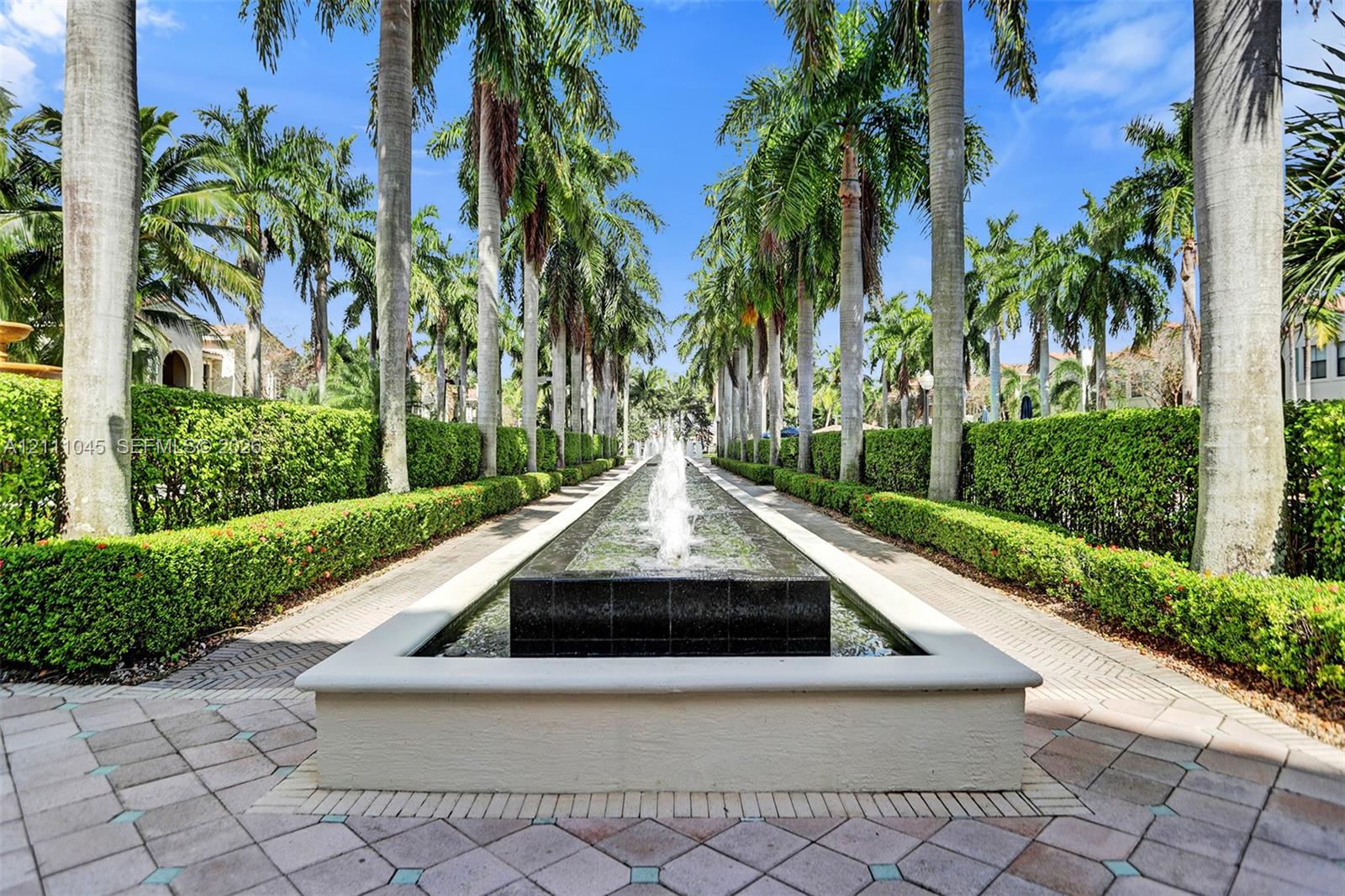 Linear water feature with a central fountain, bordered by manicured hedges, towering palm trees, and decorative paver hardscaping