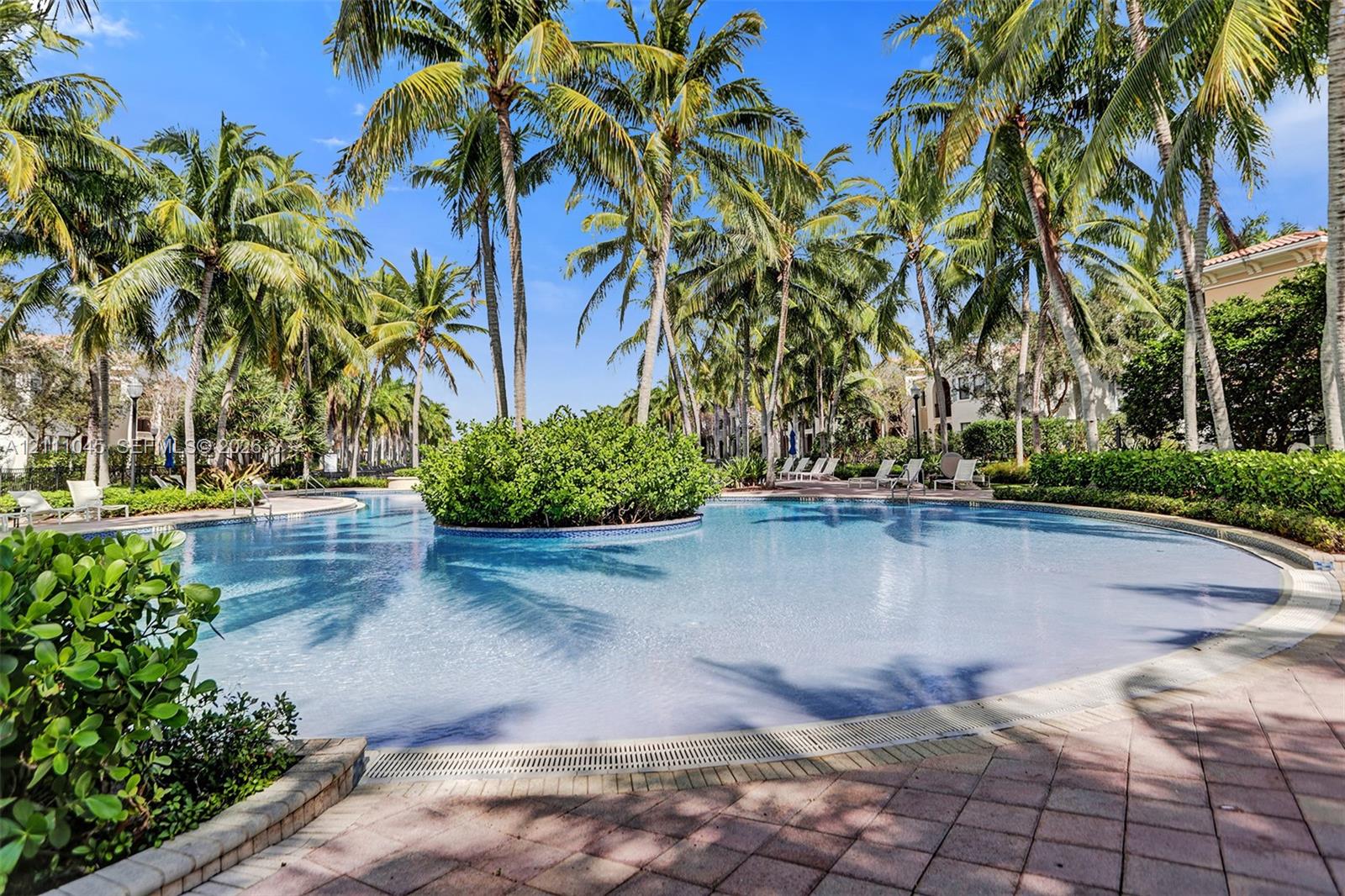 Curved swimming pool featuring a central island landscape, stone-paver decking, and mature palm tree canopy