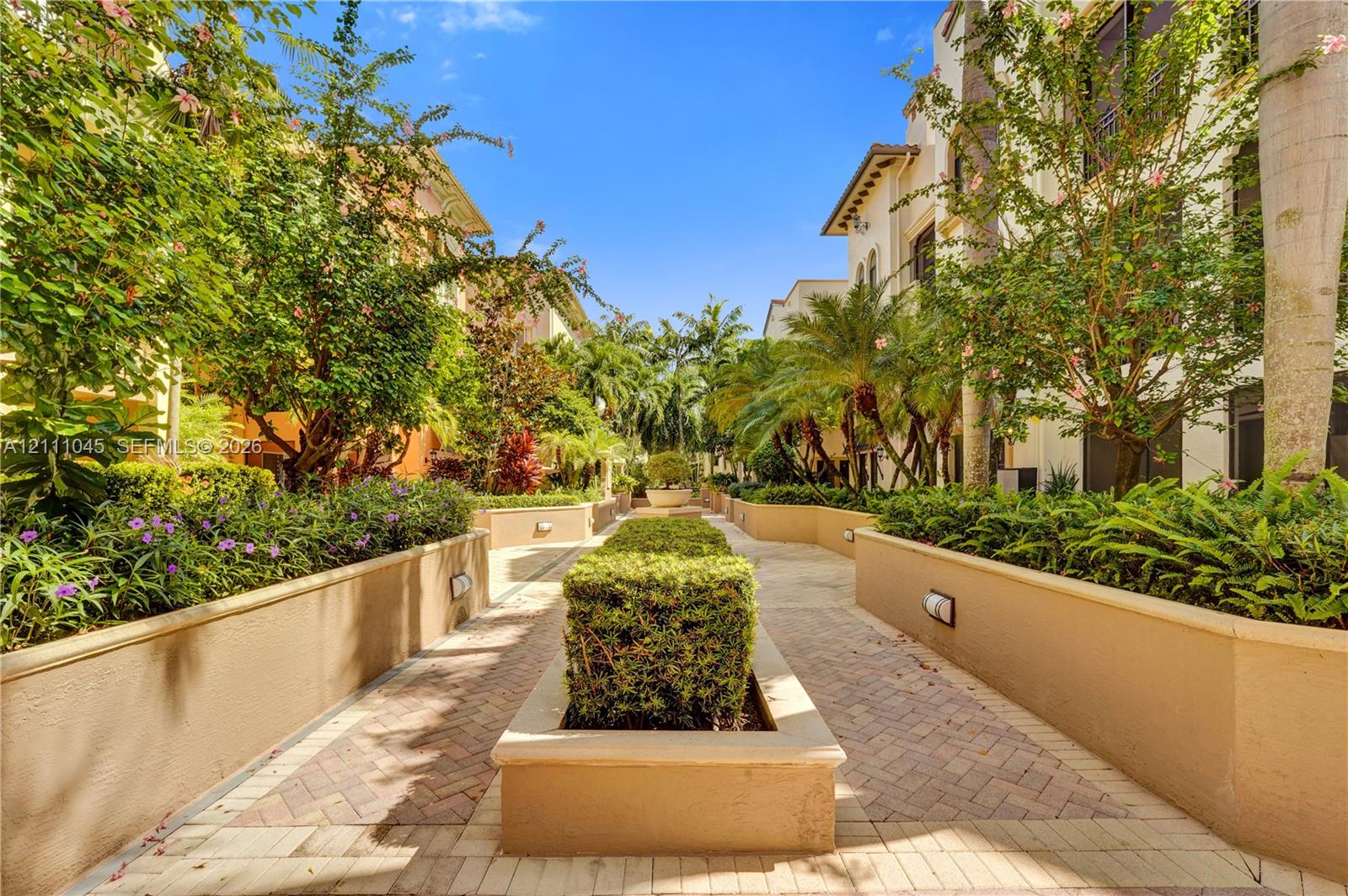 Paver walkway lined with stucco planters, manicured hedges, mature palm trees, and integrated wall lighting