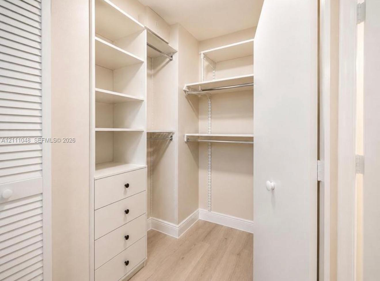 Walk-in closet featuring custom built-in shelving, integrated four-drawer storage, polished metal hanging rods, wood-finish flooring, and a white louvered door Walk-in closet featuring custom built-in shelving, integrated four-drawer storage, polished metal hanging rods, wood-finish flooring, and a white louvered door